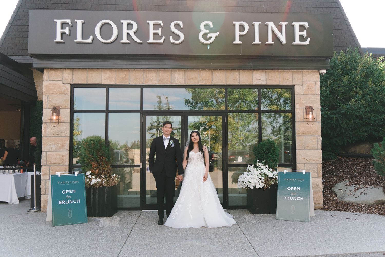 Bride and groom pose in front of Flores and Pine restaurant entrance with white floral decor.