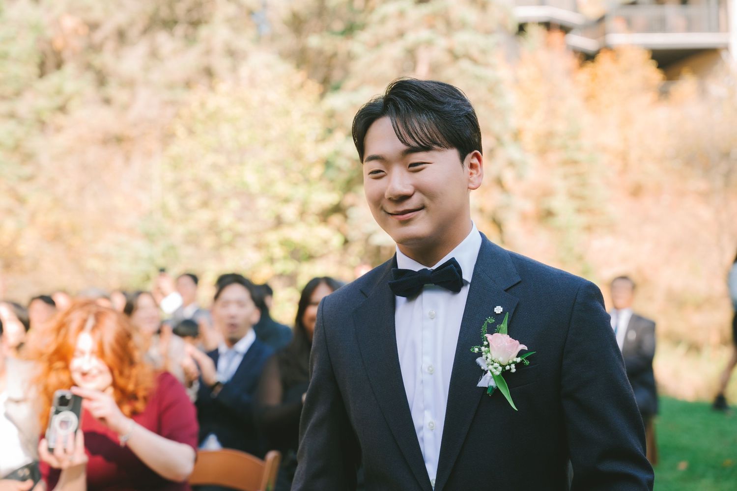 Smiling groom in black tuxedo with boutonniere stands at outdoor fall wedding ceremony.