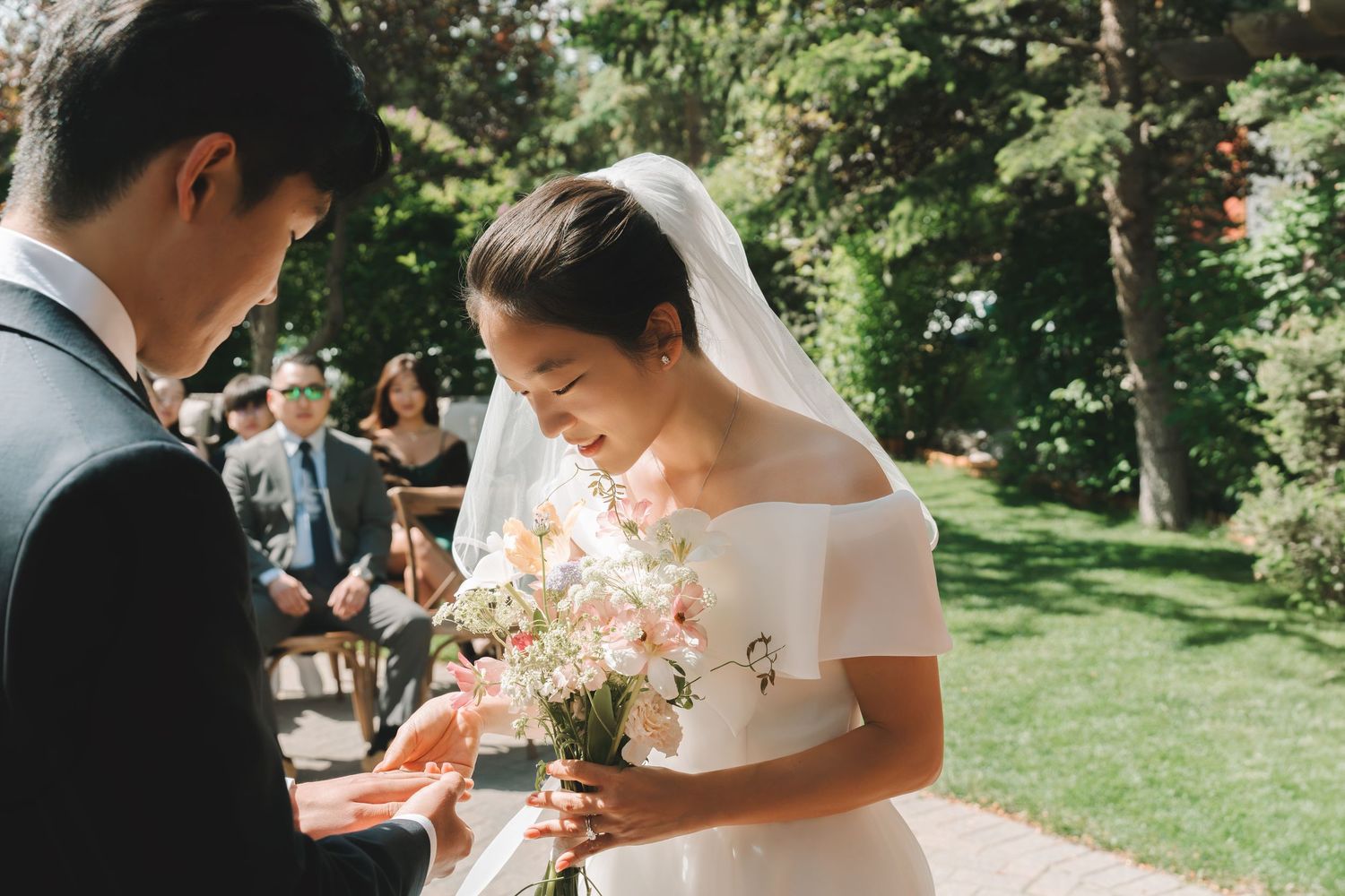 Groom places wedding ring on bride's finger as she holds a soft pink and white floral bouquet.