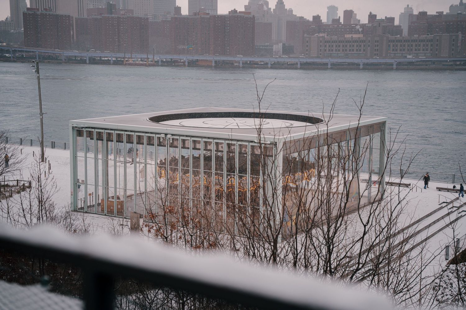Snowy winter view of a modern glass pavilion on the Hudson River waterfront with NYC skyline in the background.