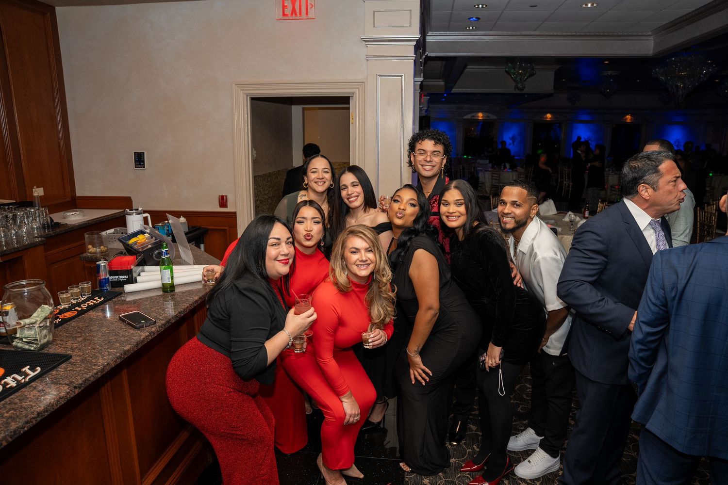 A group of friends pose and smile together near a bar at an elegant wedding reception with blue ambient lighting.