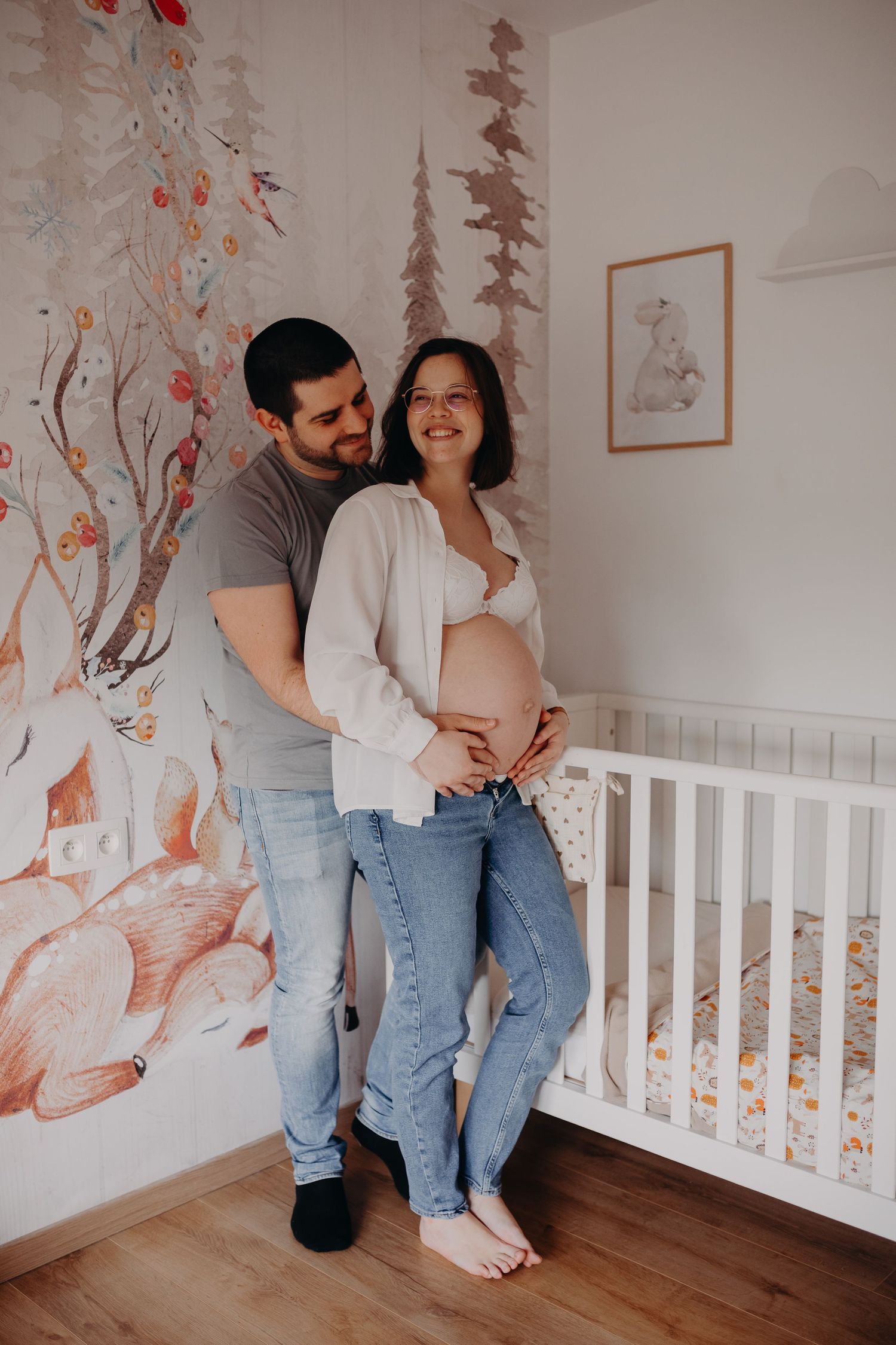 Couple in nursery room with woodland wallpaper, pregnant woman cradling baby bump, standing beside white crib.