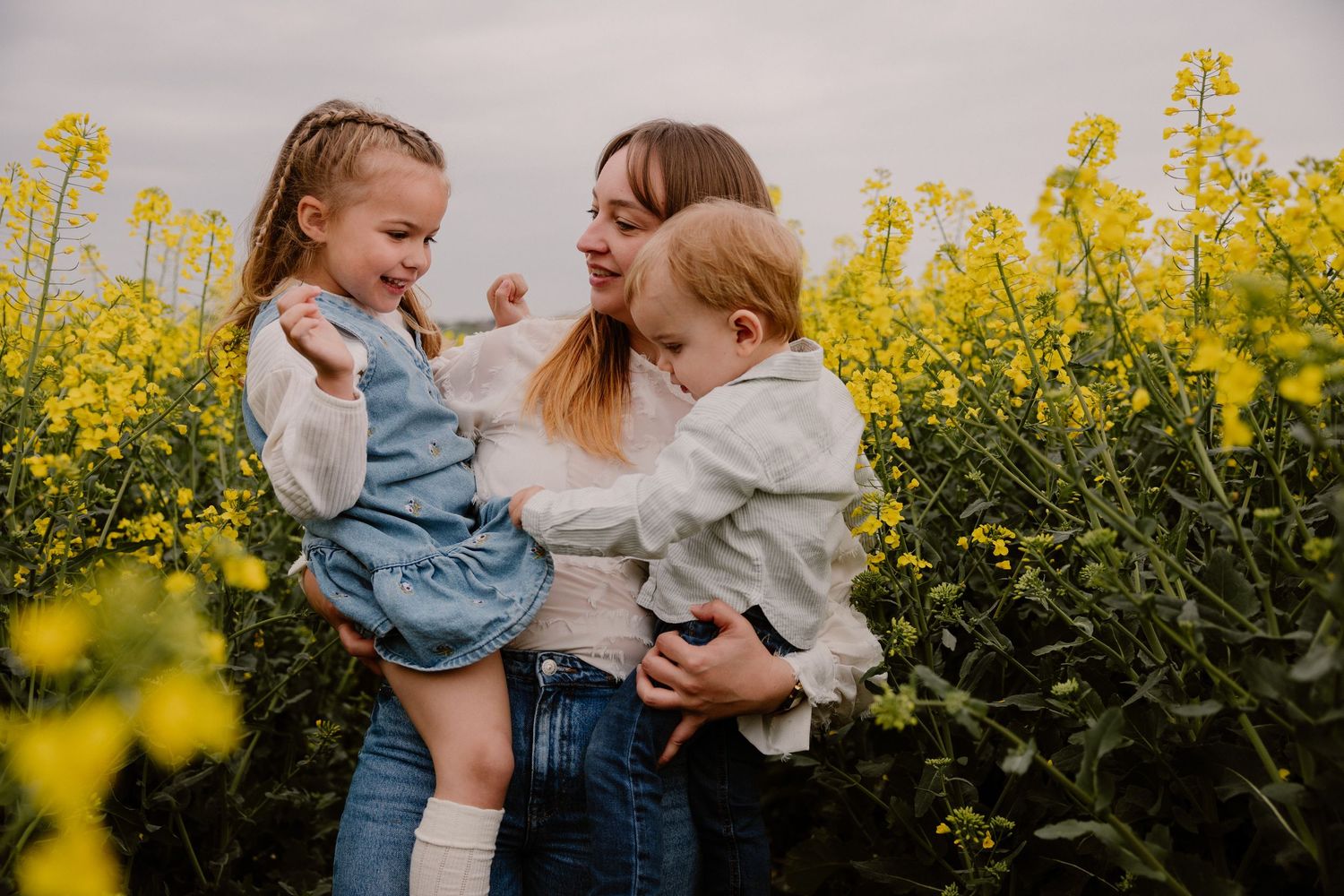 A mother holds two young children in a vibrant yellow rapeseed field, sharing a joyful moment together.