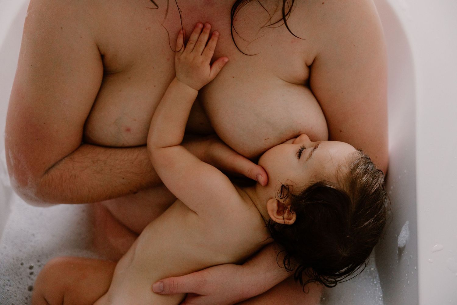 A parent tenderly holds a sleeping baby while bathing together in a white bathtub.
