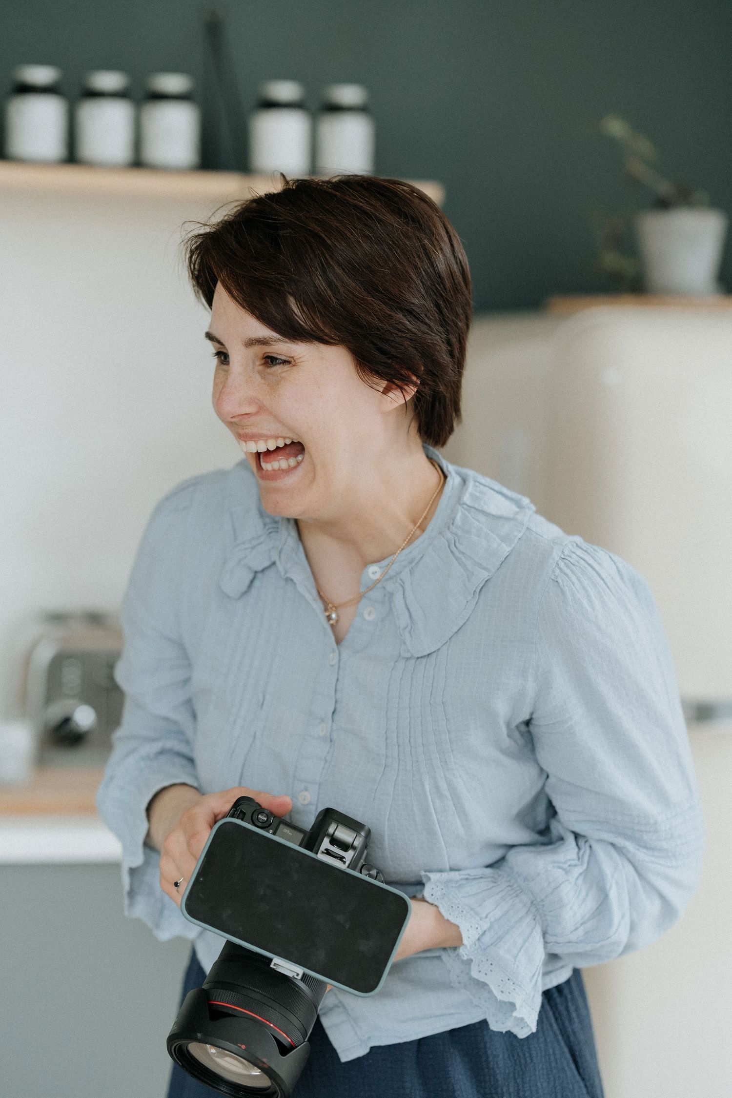 Smiling woman with short dark hair holding a camera in a bright kitchen setting.