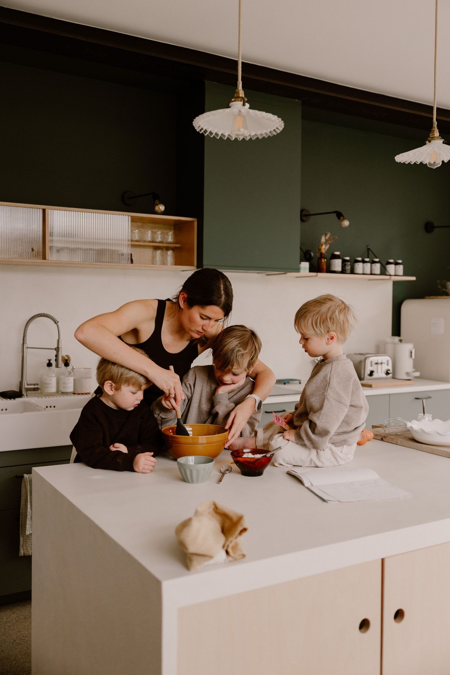 A mother and three young children cooking together at a modern kitchen island with green cabinetry and pendant lights.