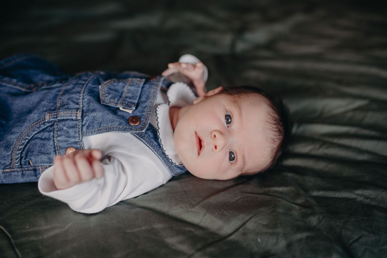 Newborn baby in denim overalls lying on dark surface, looking up with bright blue eyes.