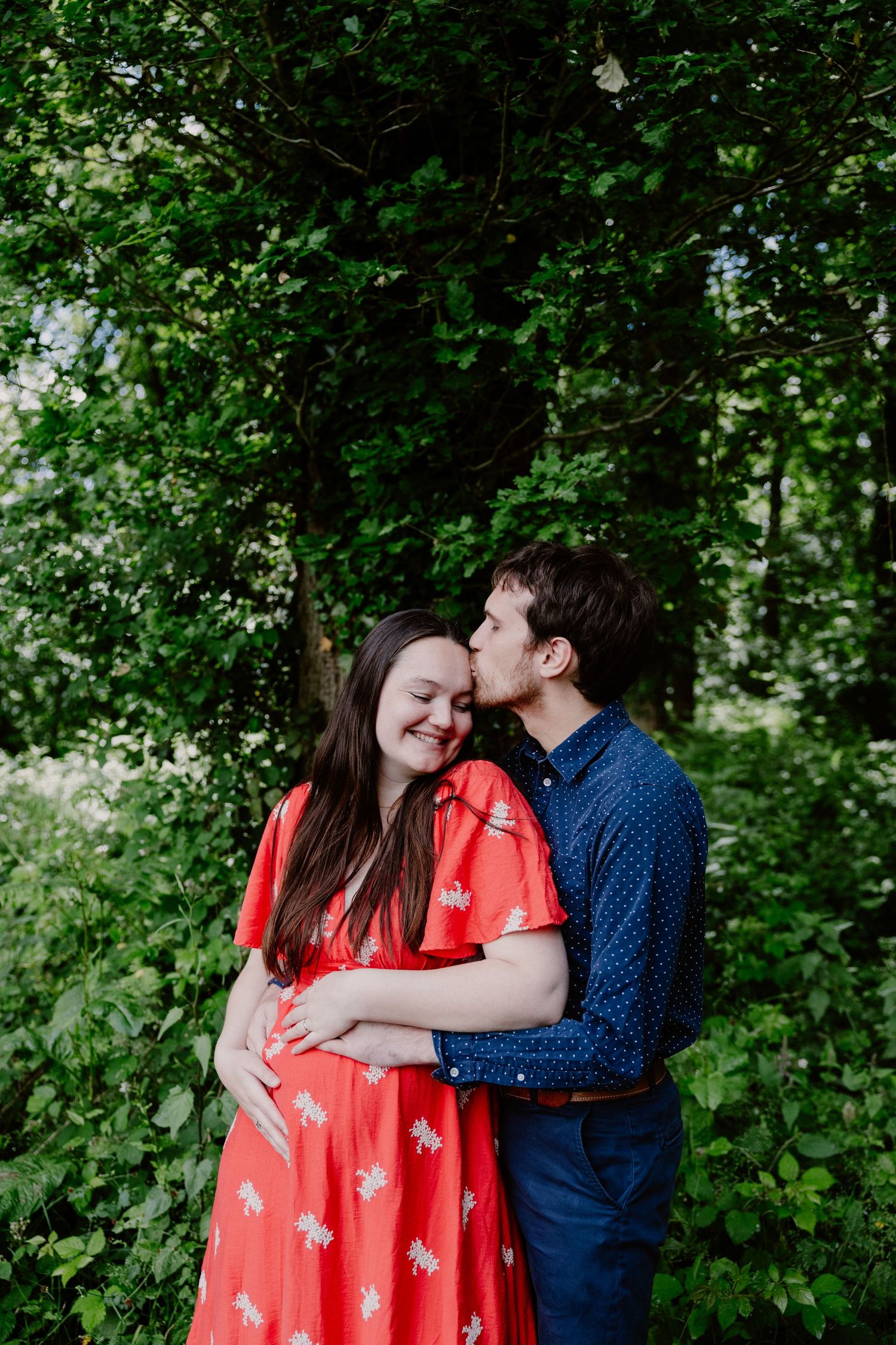 A couple shares a tender kiss outdoors, woman in red floral dress cradling baby bump, surrounded by lush green foliage.