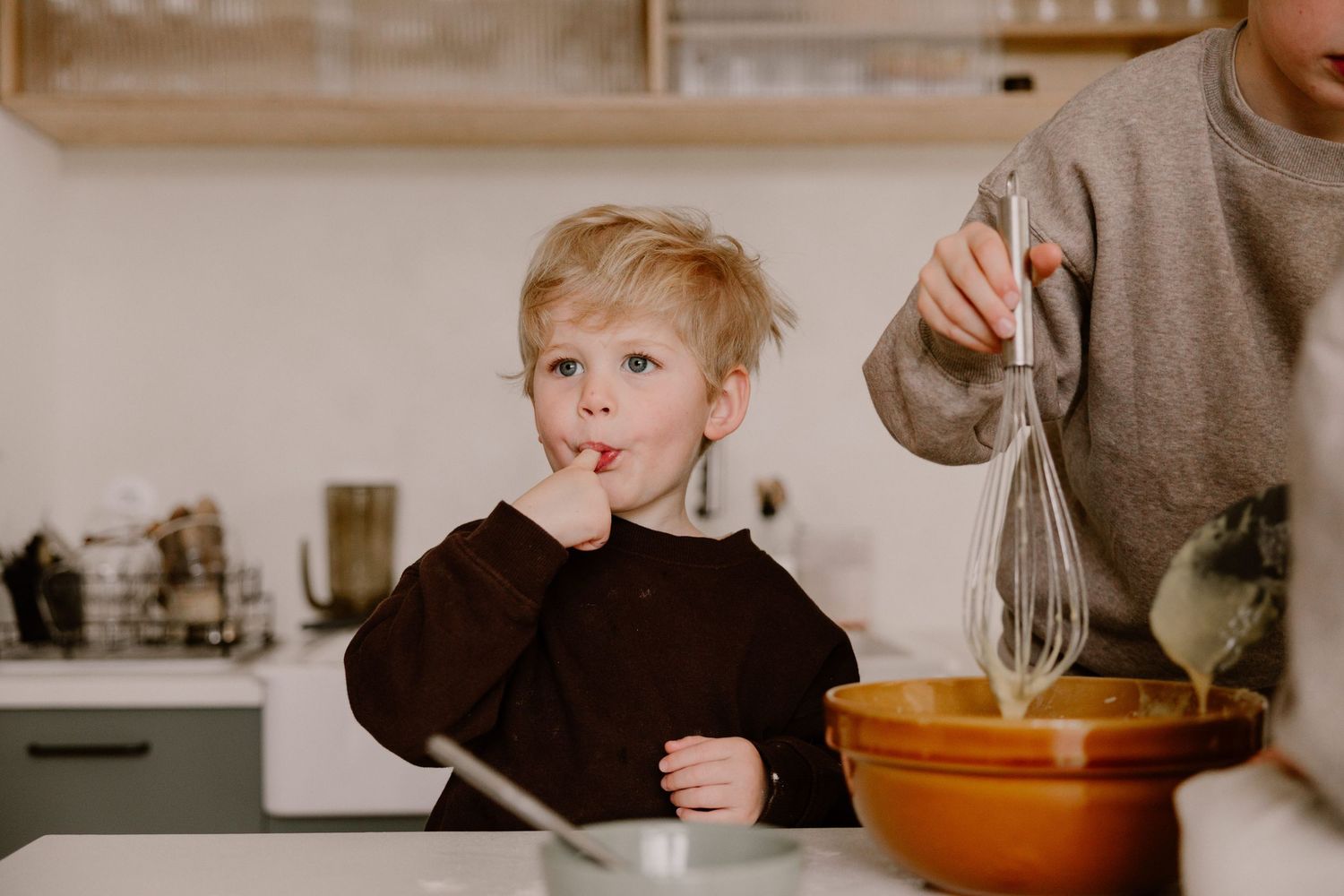 A young blonde boy licks his fingers while helping bake in a kitchen, as an adult whisks batter in a bowl.