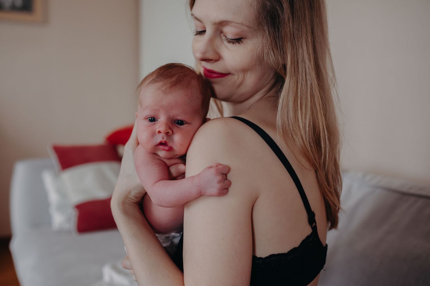 A mother with red lips holds her alert newborn baby against her shoulder at home.