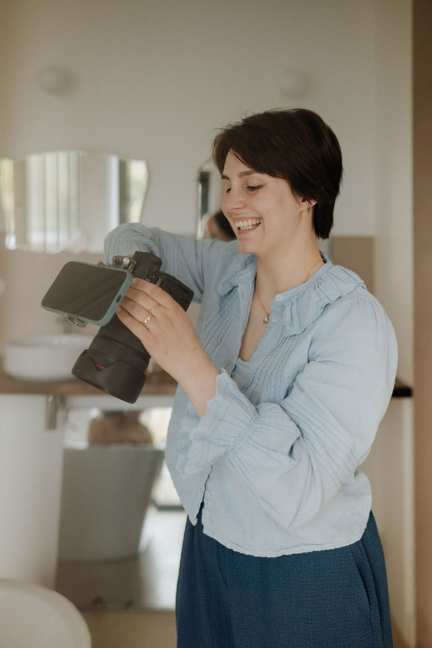 Smiling woman with short dark hair holds DSLR camera indoors, wearing blue linen shirt in bright room.