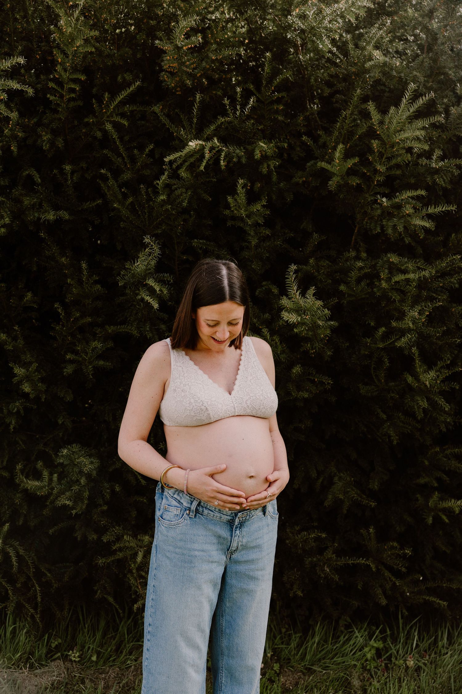 Pregnant woman in lace bralette and jeans cradling baby bump outdoors near green ferns.