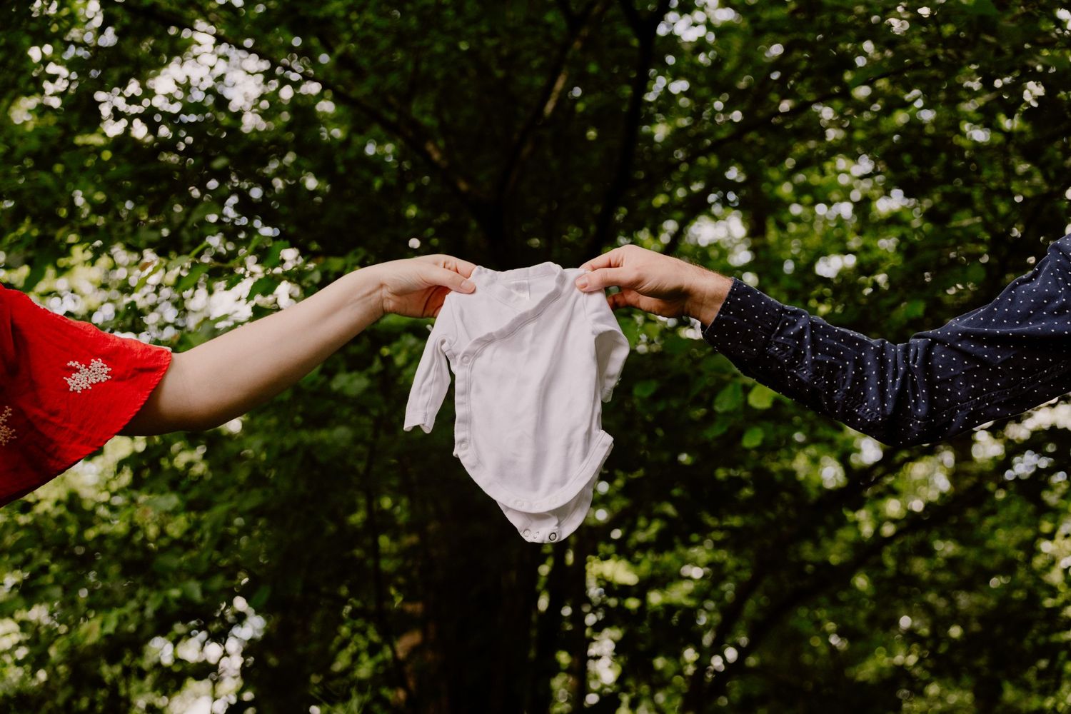 Two people holding a tiny white baby onesie outdoors surrounded by lush green trees, announcing a pregnancy.