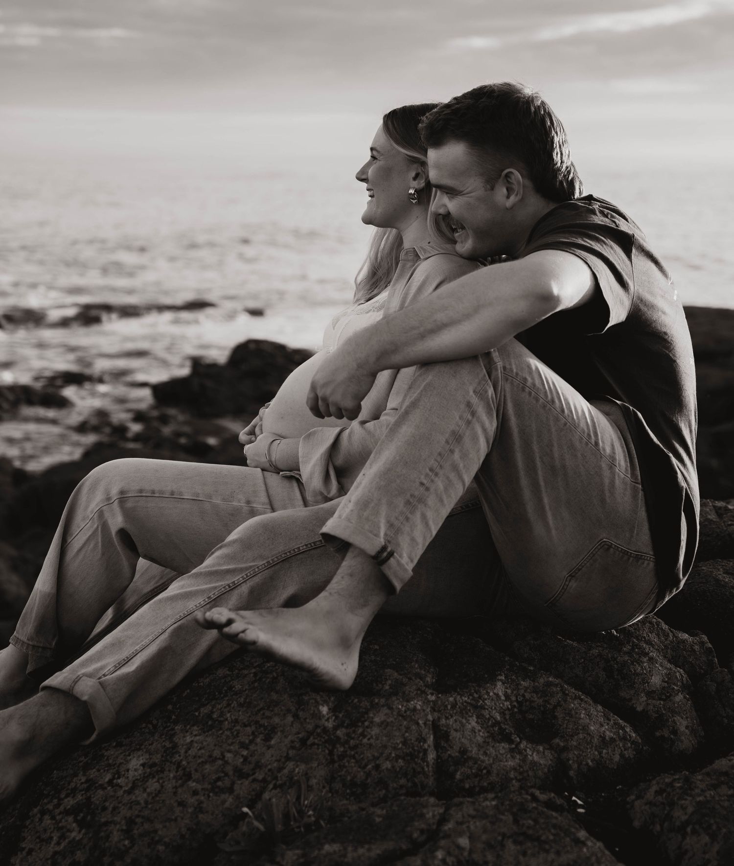 Couple sitting on coastal rocks in black and white, embracing romantically with ocean waves in the background.