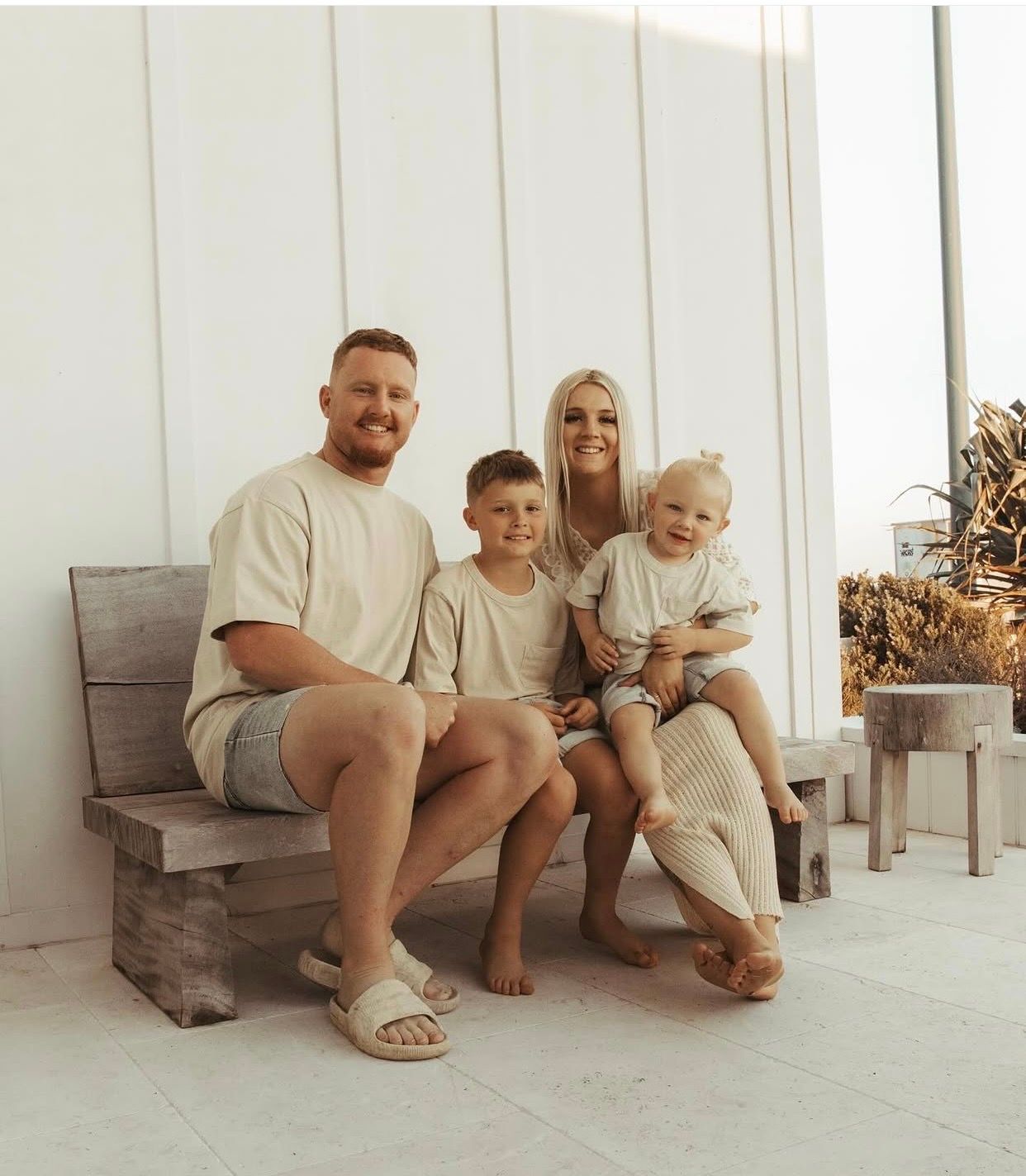 Family of four in matching neutral outfits smiling together on a rustic wooden bench indoors.