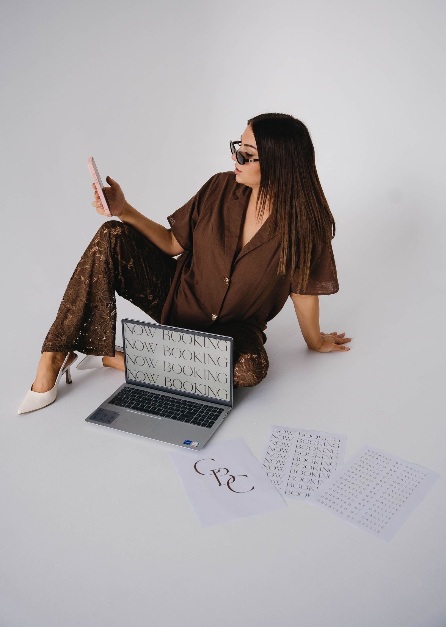 Woman in brown outfit sitting on floor with laptop displaying 'Now Booking' text and papers nearby.