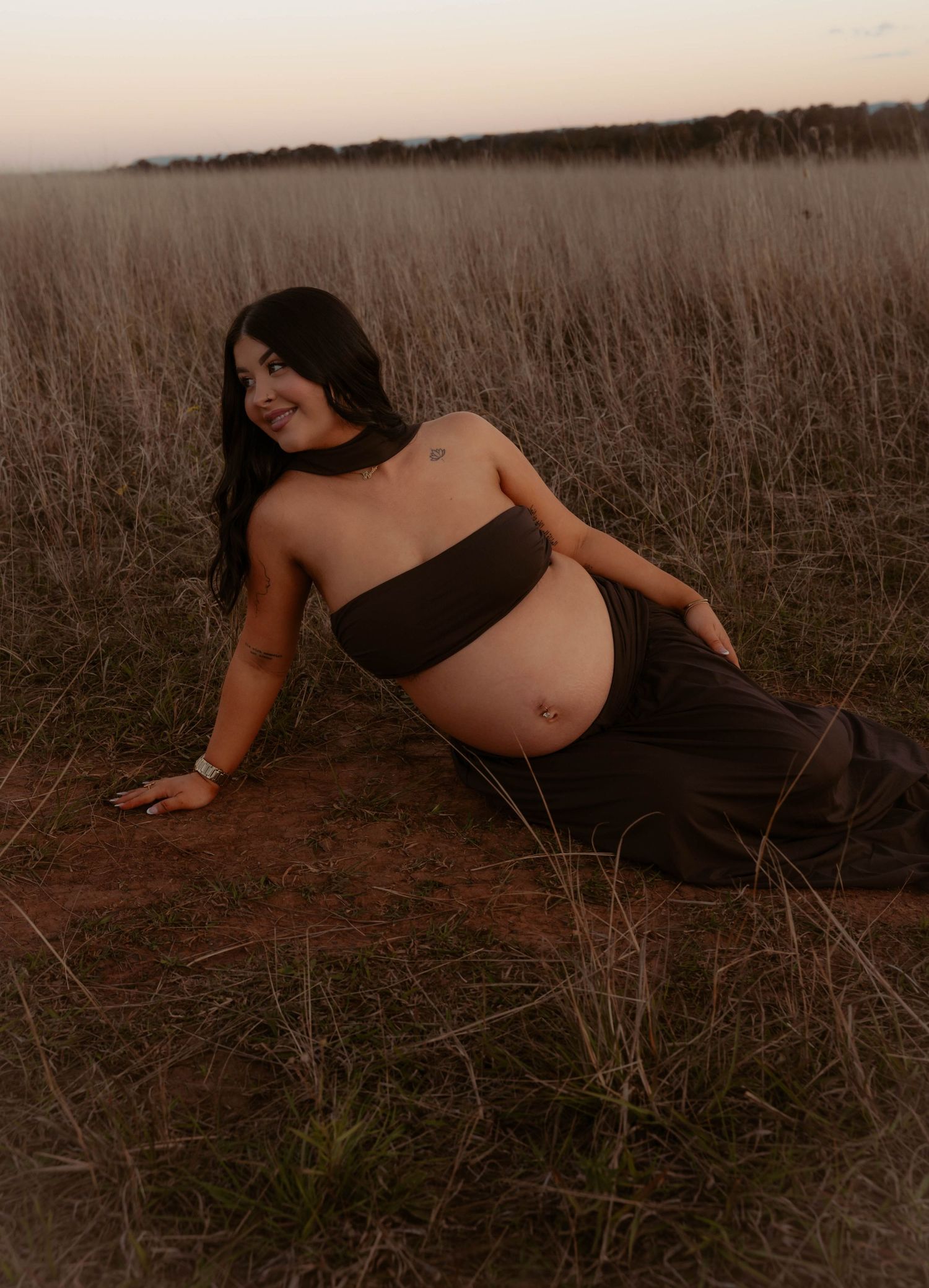 Pregnant woman posing in a golden field at dusk, wearing a dark bandeau top and maxi skirt.