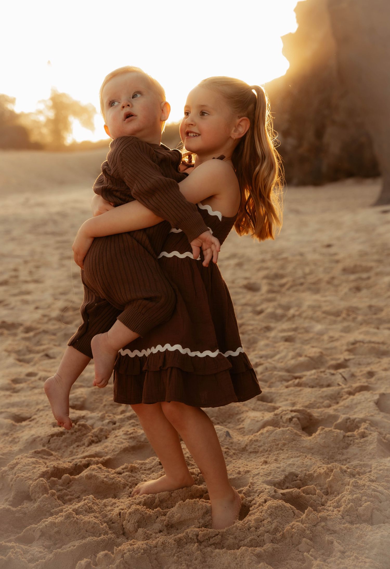 A young girl holds a baby sibling on a sandy beach at golden hour in matching brown dresses.