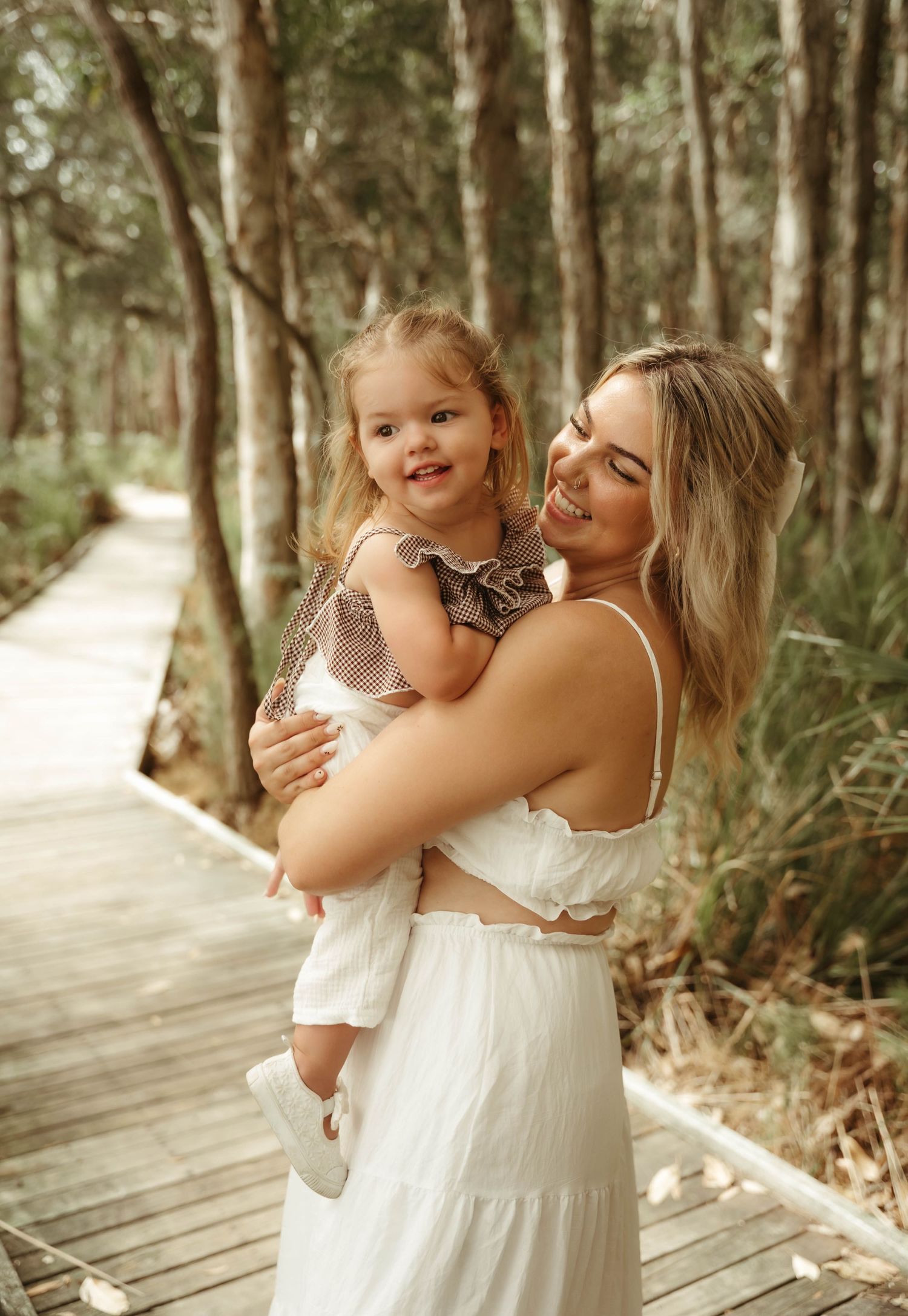 Smiling mother in white dress holds young daughter on a scenic wooden boardwalk in the forest.
