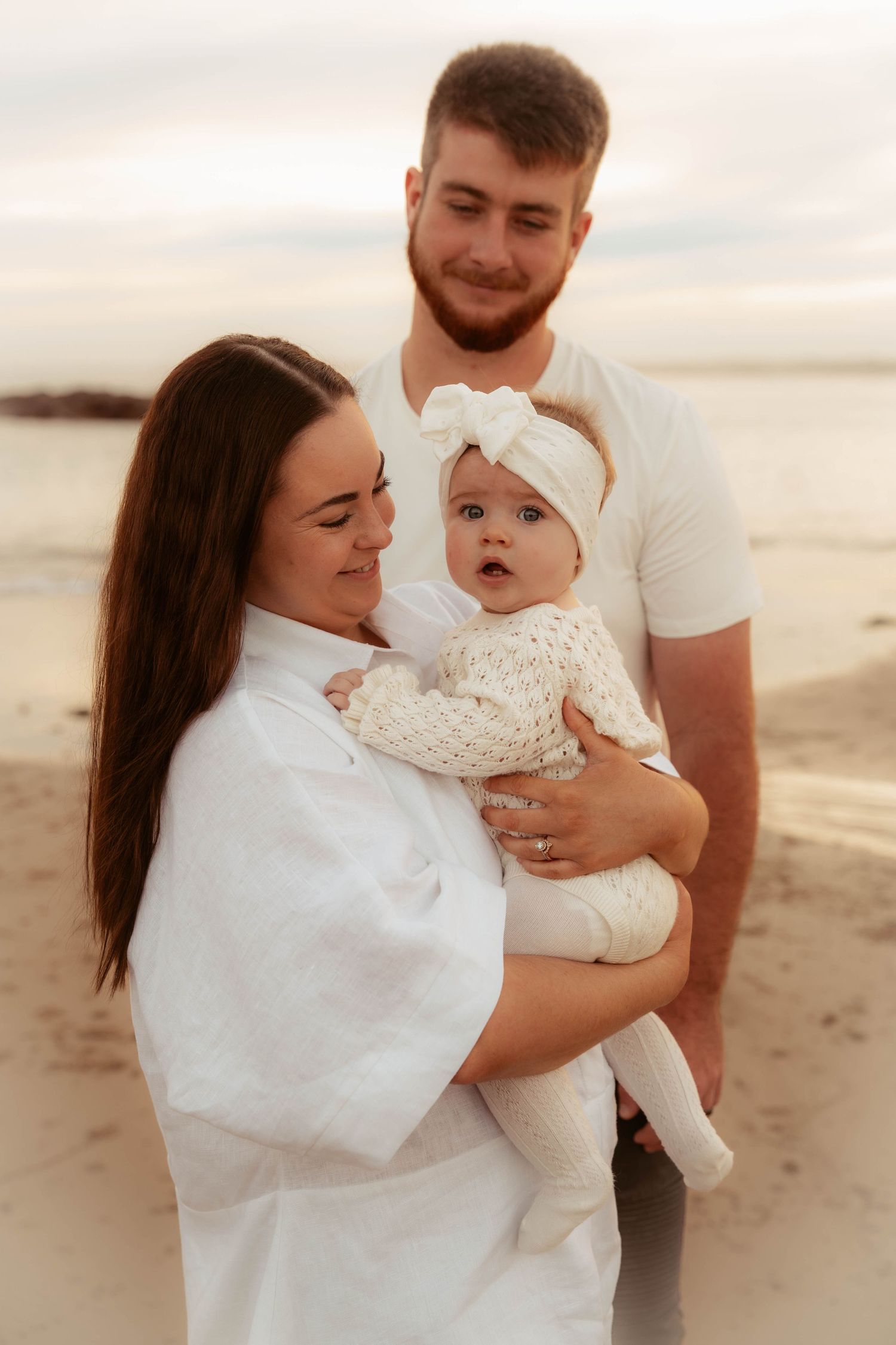 Young couple holding their baby girl in white bow headband on a serene beach at dusk.