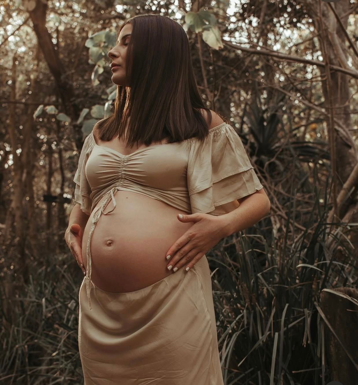 Pregnant woman in gold maternity dress cradling baby bump outdoors among lush green foliage.