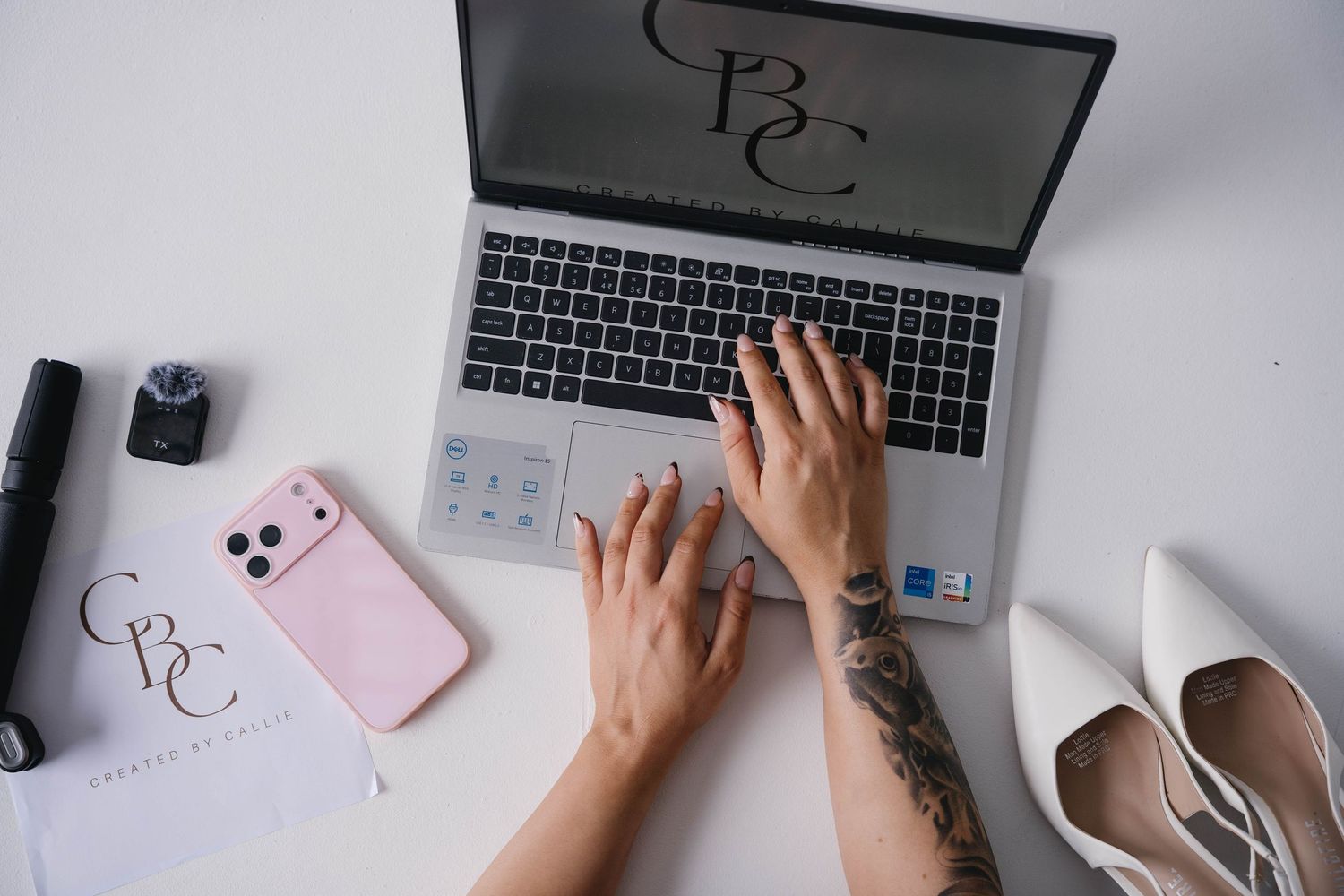 Tattooed hands typing on a laptop with a pink phone case, white heels, and branded stationery on a white desk.