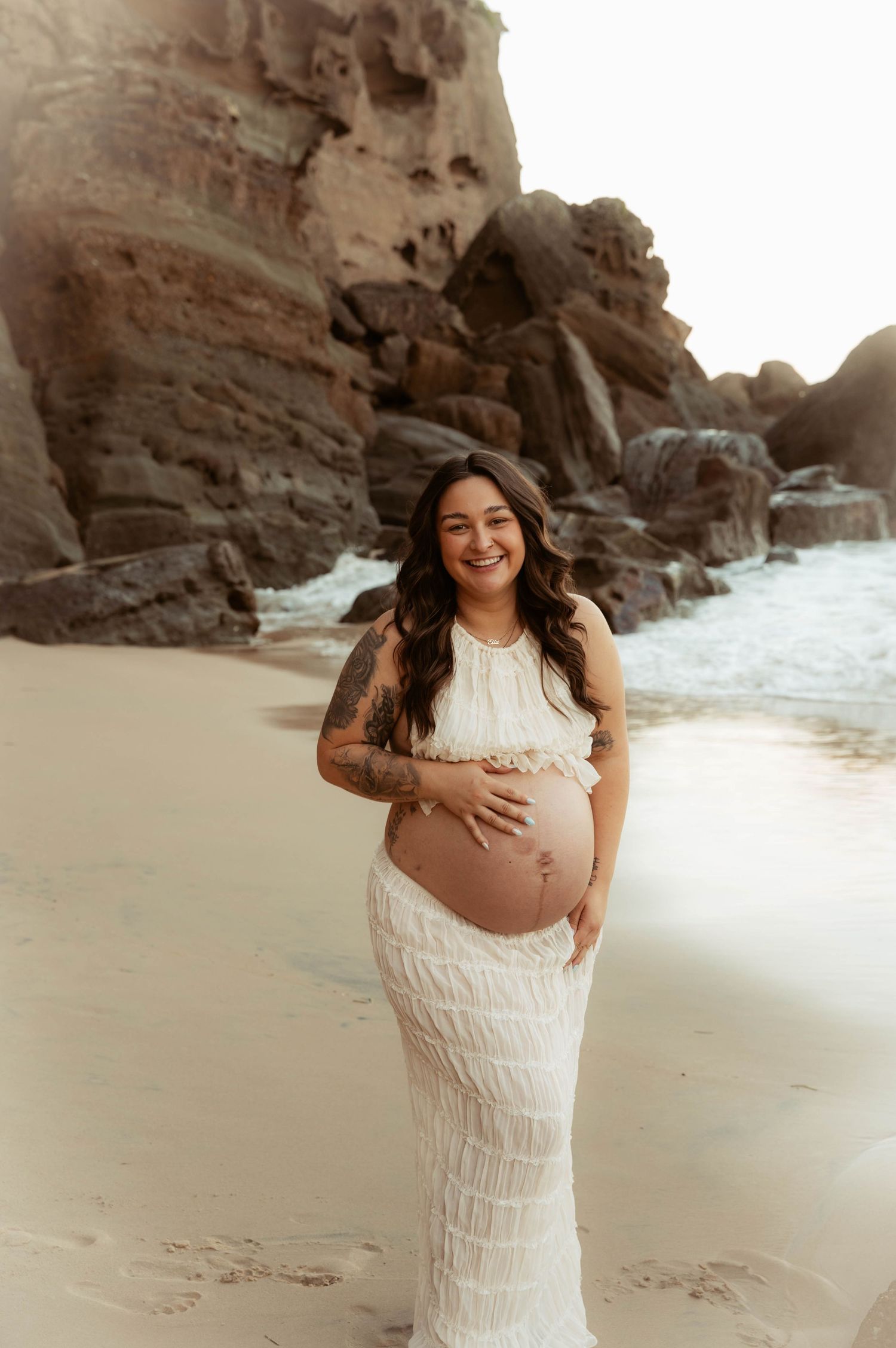 Smiling pregnant woman in white lace dress cradling baby bump on sandy beach with rocky cliffs.