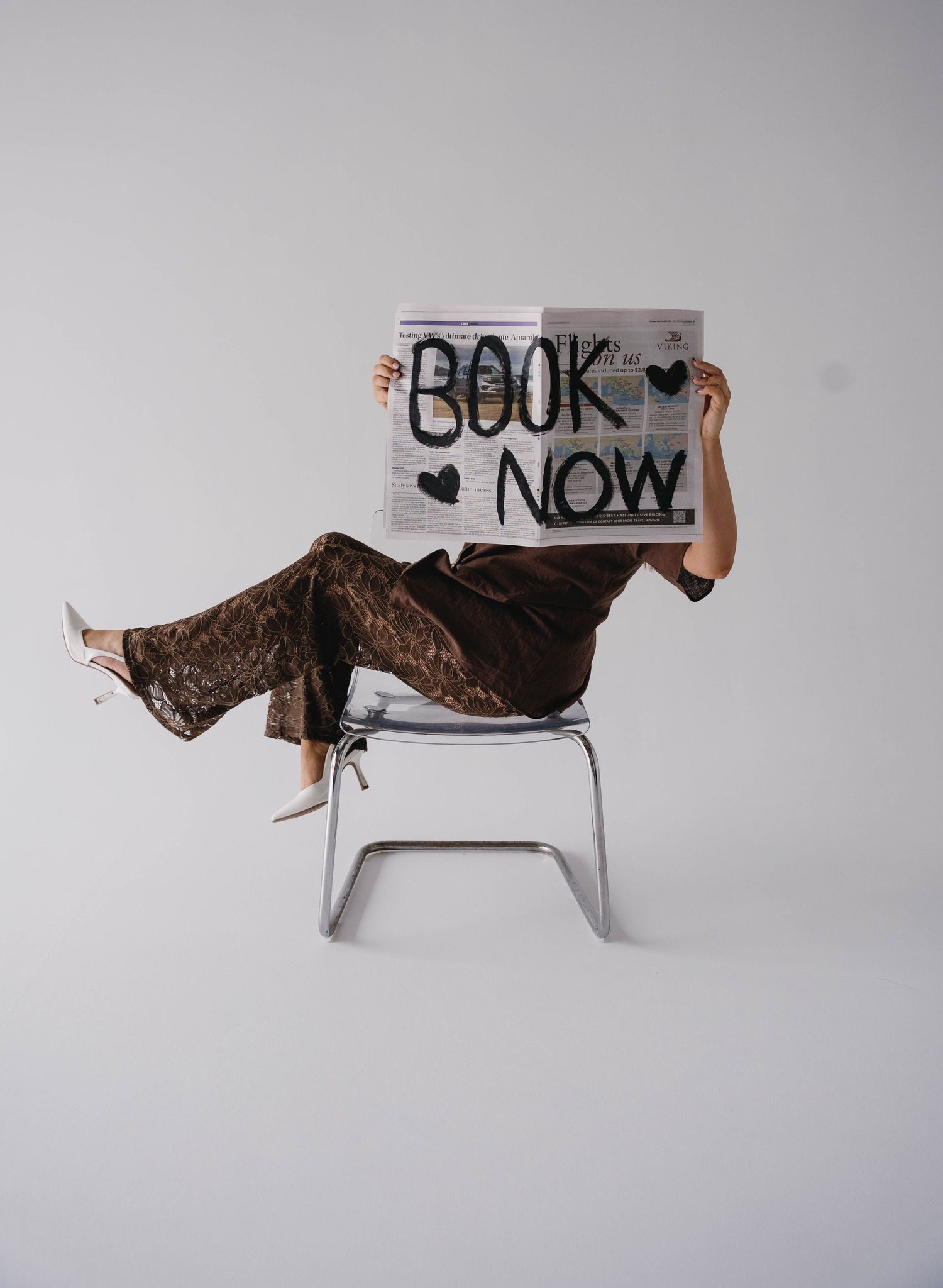 Person sitting on a modern chair holding a newspaper sign reading Book Now with heart decorations.