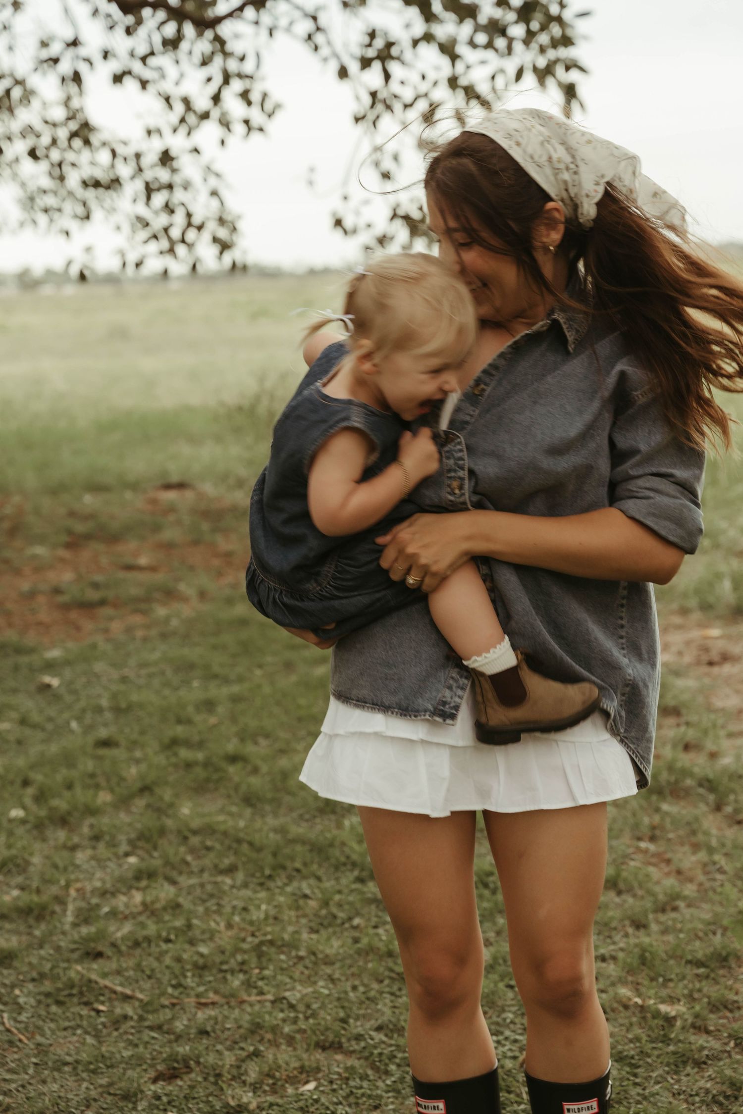 Loving mother cradling sleeping toddler outdoors in lush green field wearing denim shirt and white skirt.