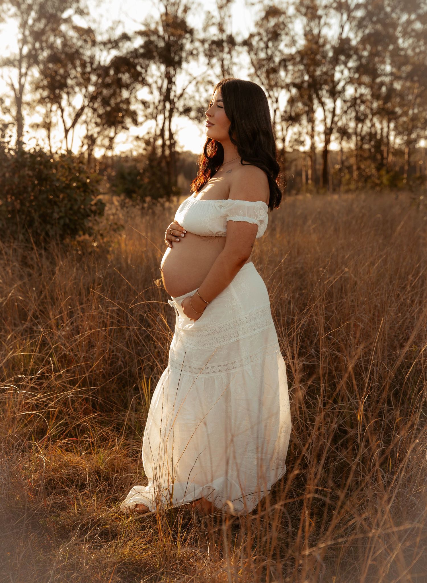Pregnant woman in white off-shoulder lace gown standing in golden dry grass field at sunset.