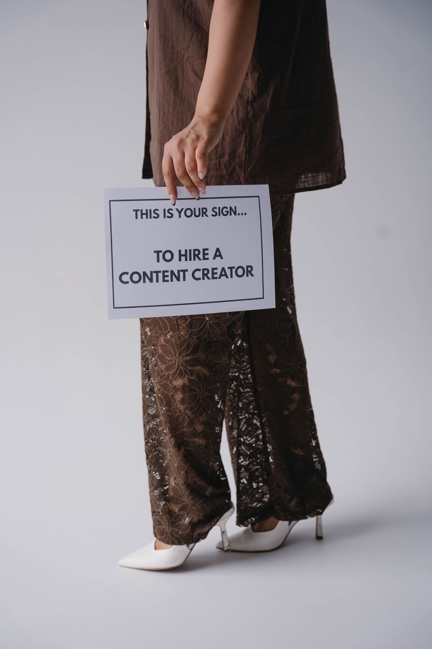 Woman in brown lace pants holding a sign reading 'This Is Your Sign To Hire A Content Creator' on white background.