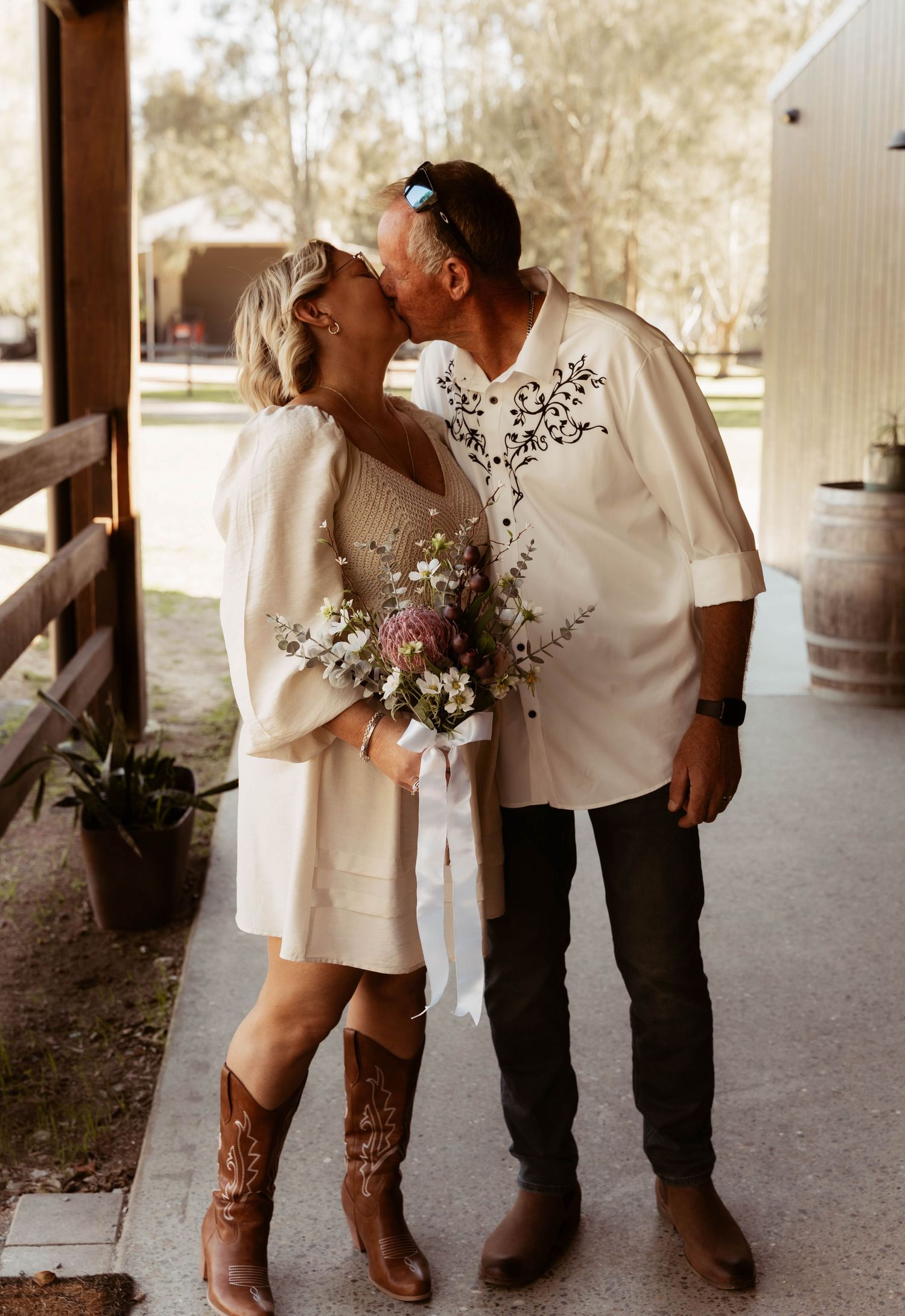 Newlywed couple kissing outdoors, bride in short white dress with cowboy boots holding wildflower bouquet.
