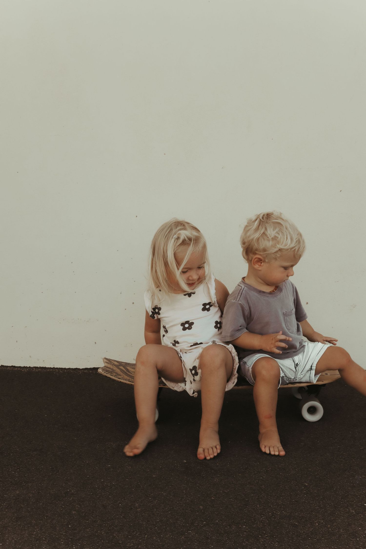 Two blonde toddlers sitting together on a skateboard against a white wall outdoors.