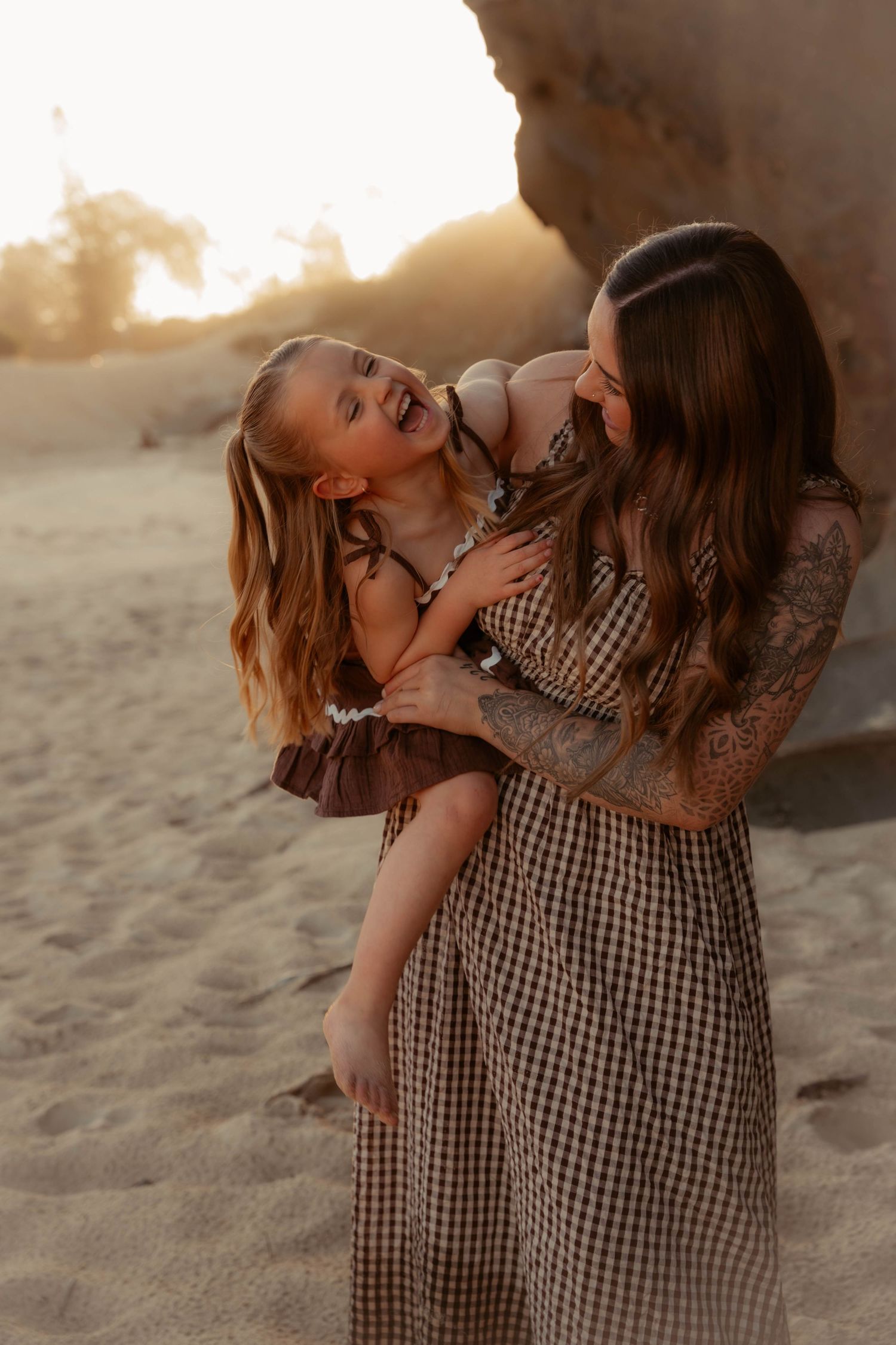 Mother holds her laughing young daughter on a sandy beach at golden hour, both wearing matching gingham dresses.