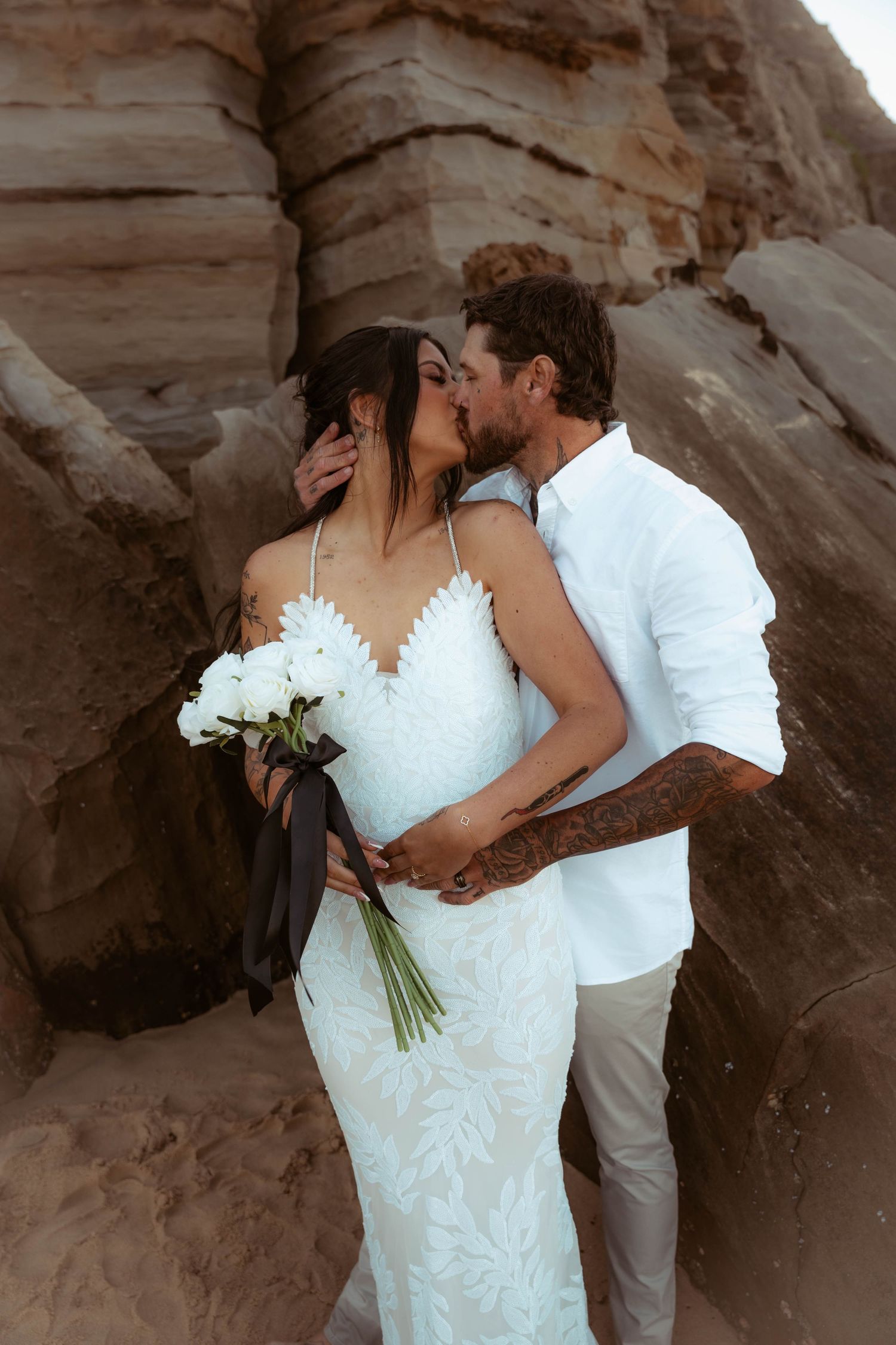 Bride and groom kissing in front of sandstone rocks, bride in lace gown holding white bouquet.