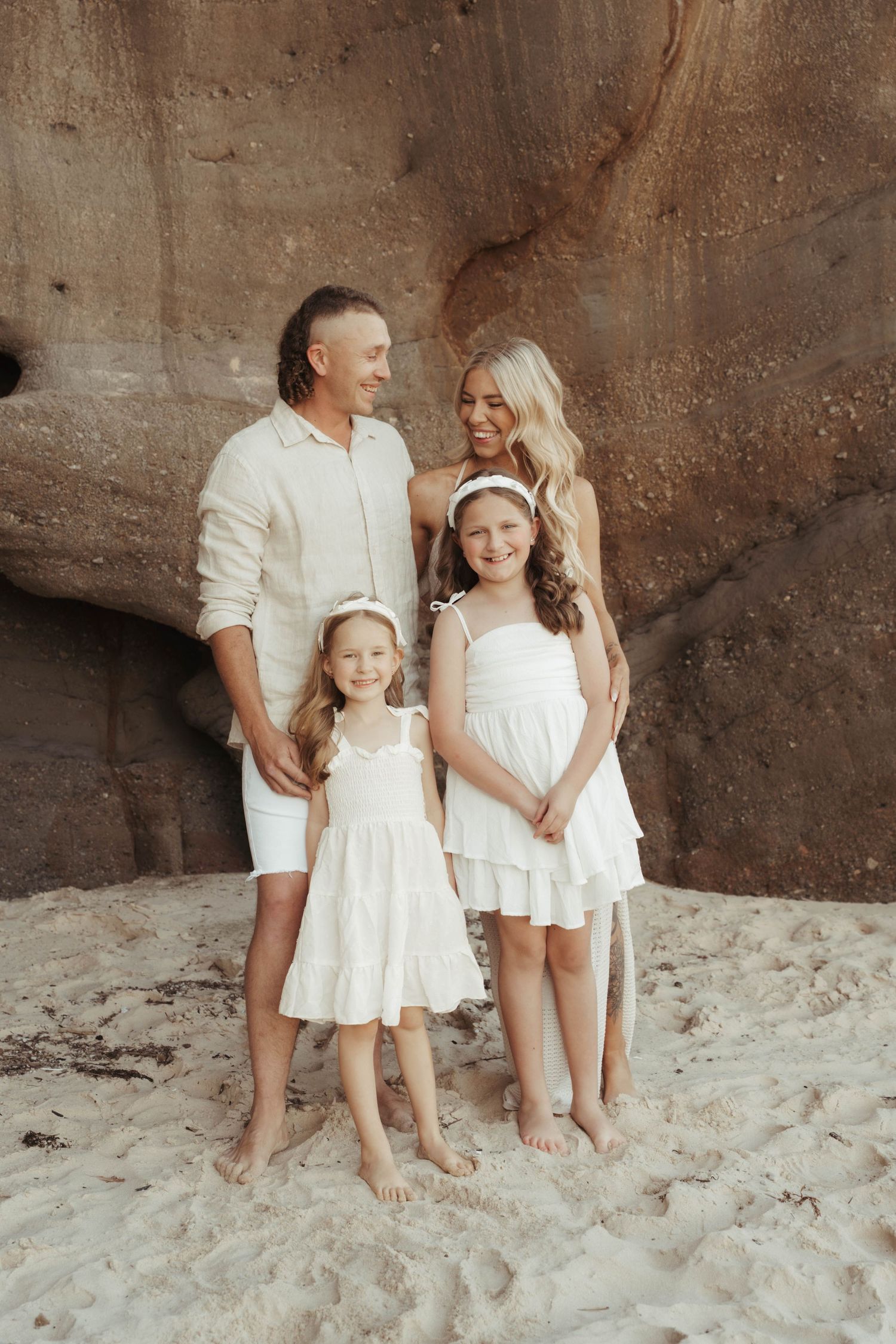Happy family of four dressed in white posing barefoot on sandy beach against rock formation.