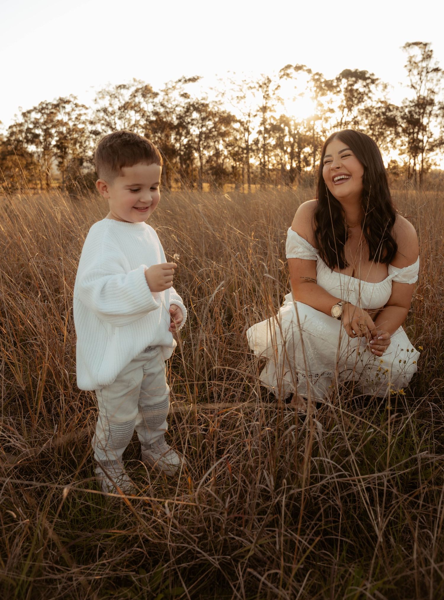 Mother in white dress kneels laughing beside her young son in a golden grass field at sunset.