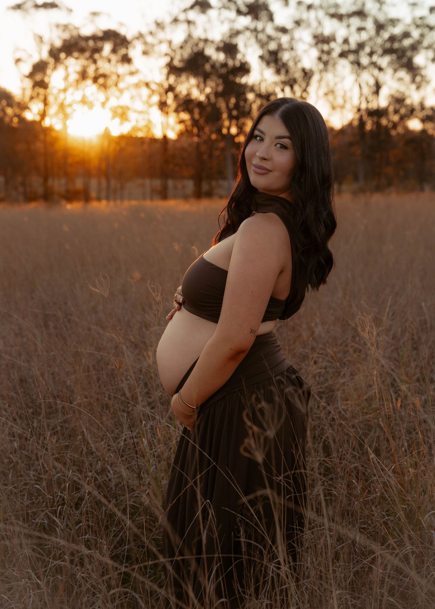 Pregnant woman in dark outfit posing in golden field at sunset showcasing baby bump.