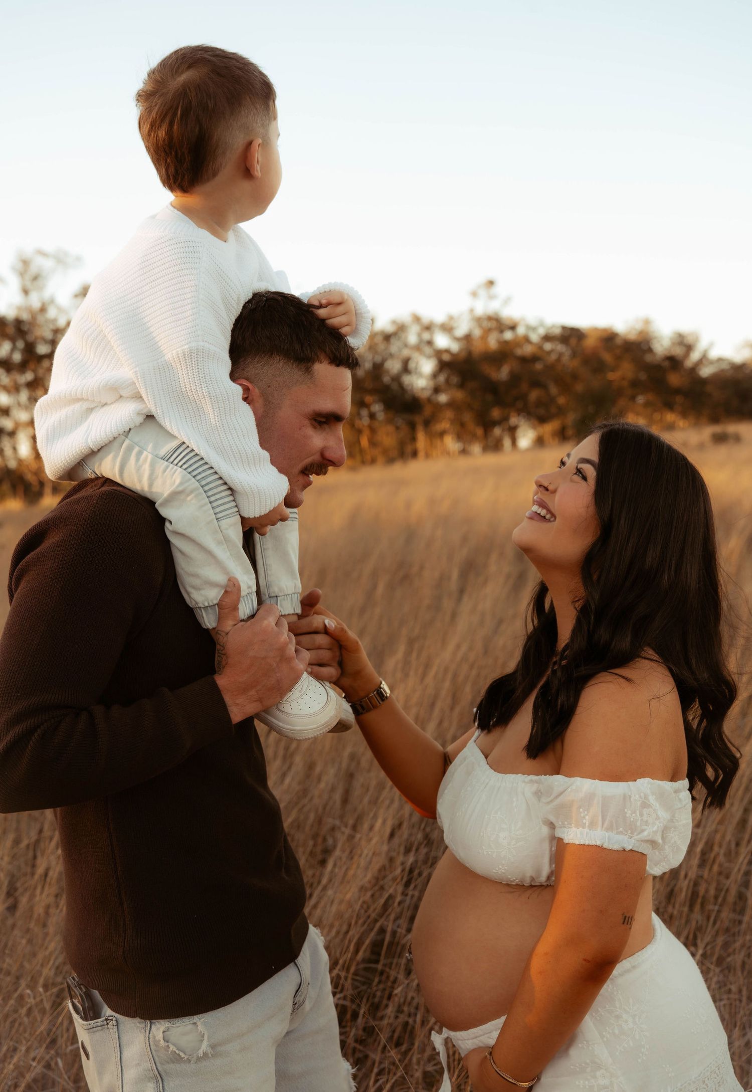 Pregnant woman in white lace dress smiling at partner holding toddler in golden field.