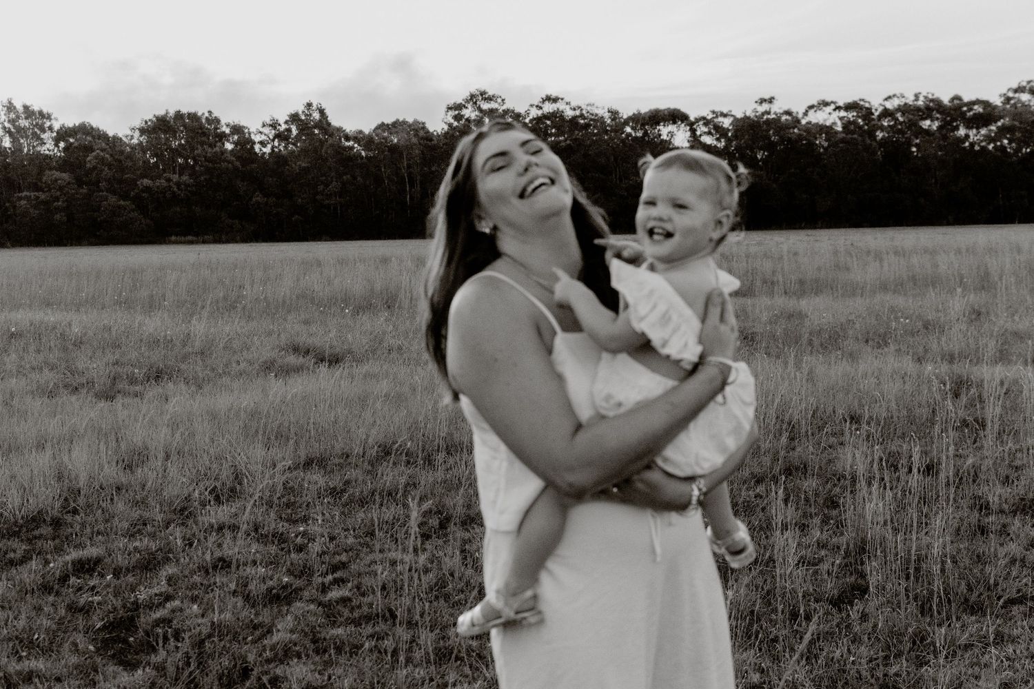 A laughing mother holds her happy baby in a black and white outdoor portrait in a grassy field.