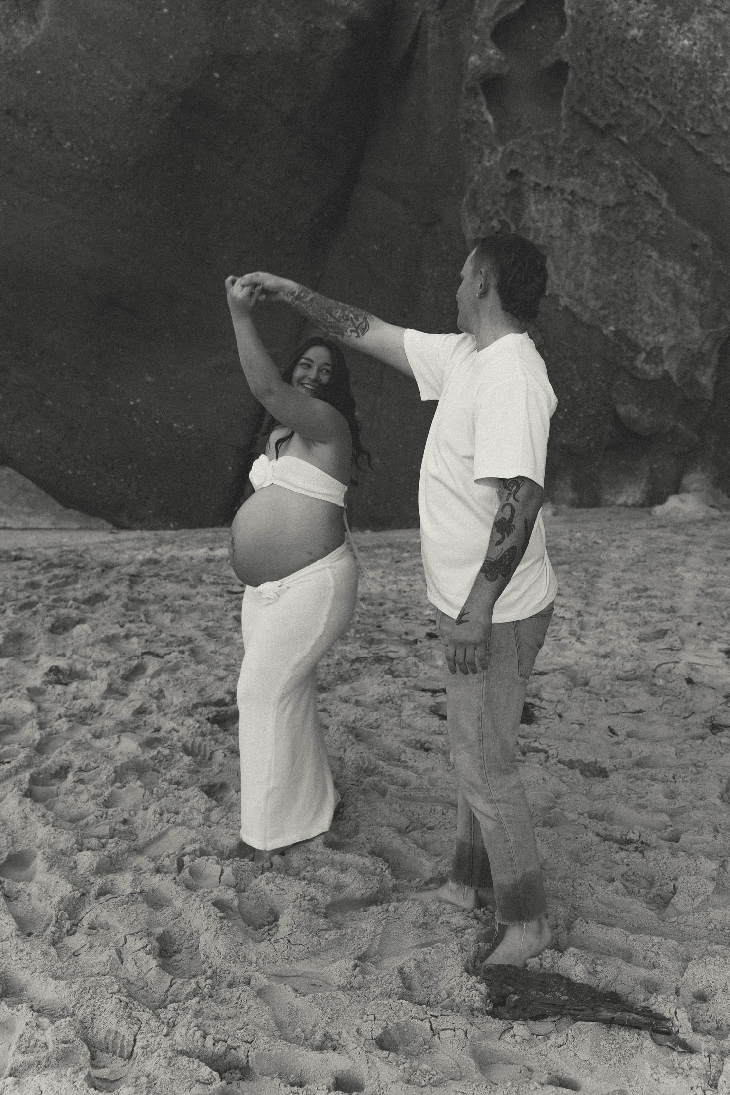 Black and white photo of expectant couple dancing on beach, man spinning pregnant woman near large rocks.