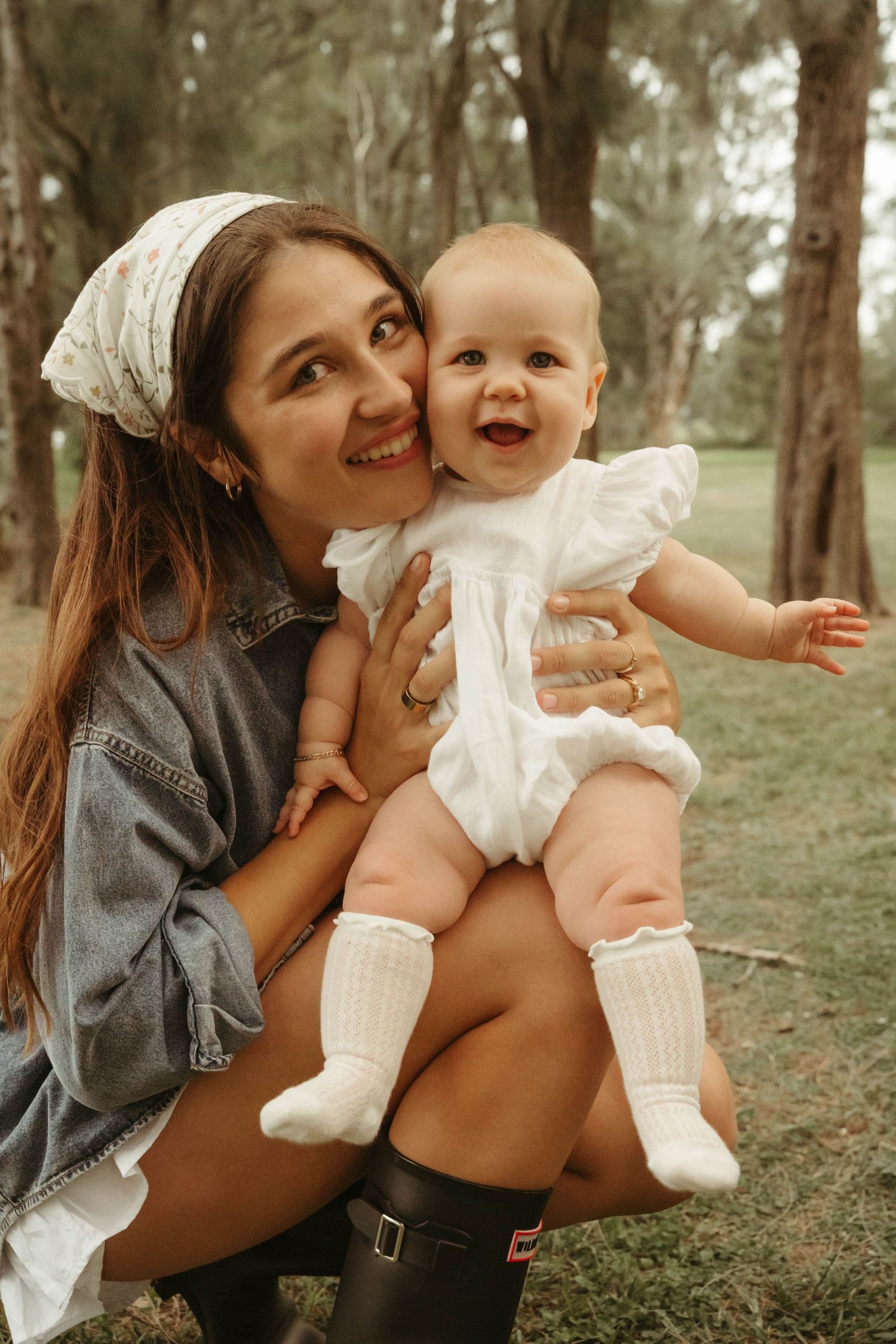 Young mother crouching in a park holding her laughing baby in a white romper and frilly socks.