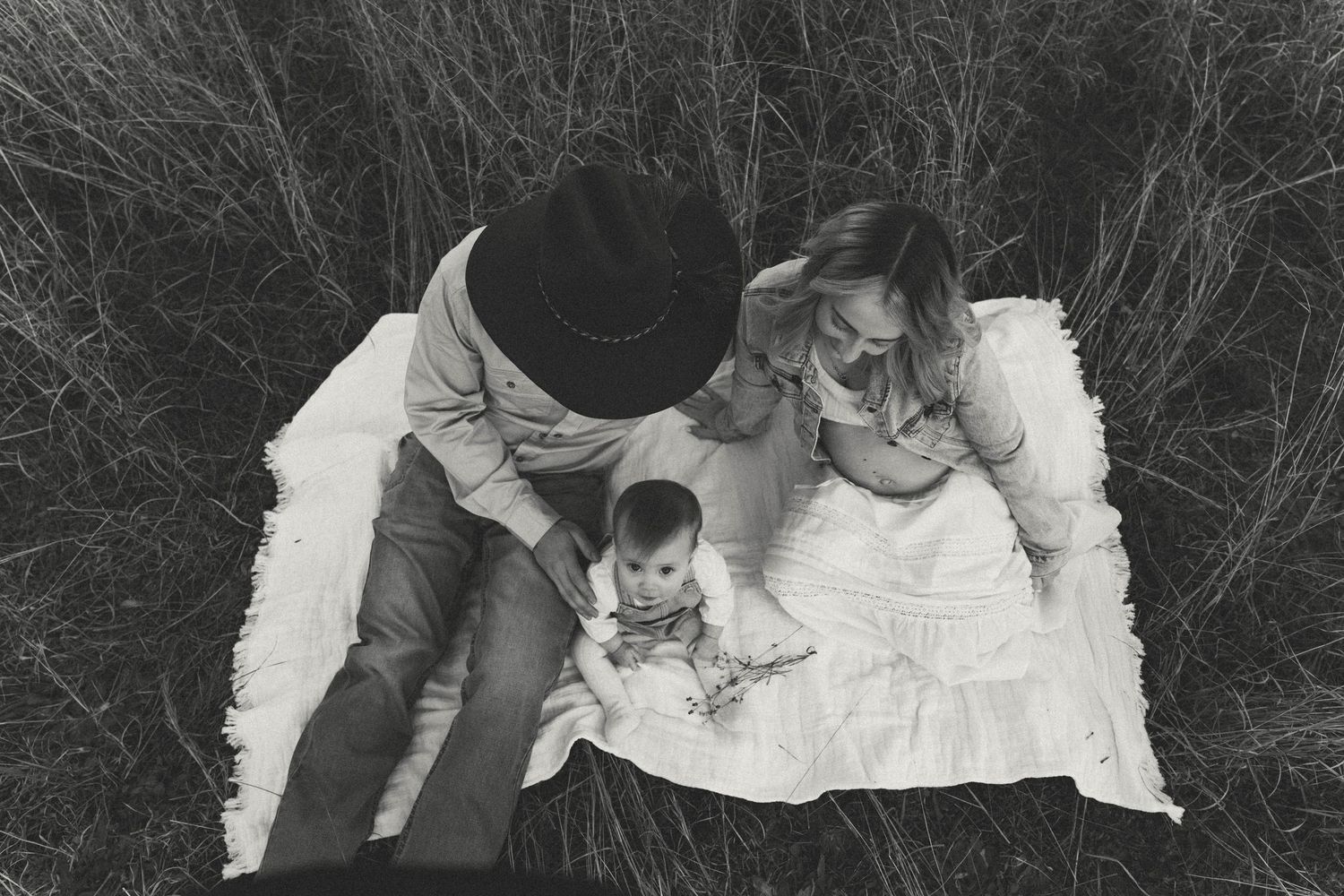 Black and white aerial photo of couple sitting on blanket with baby in tall grass field.