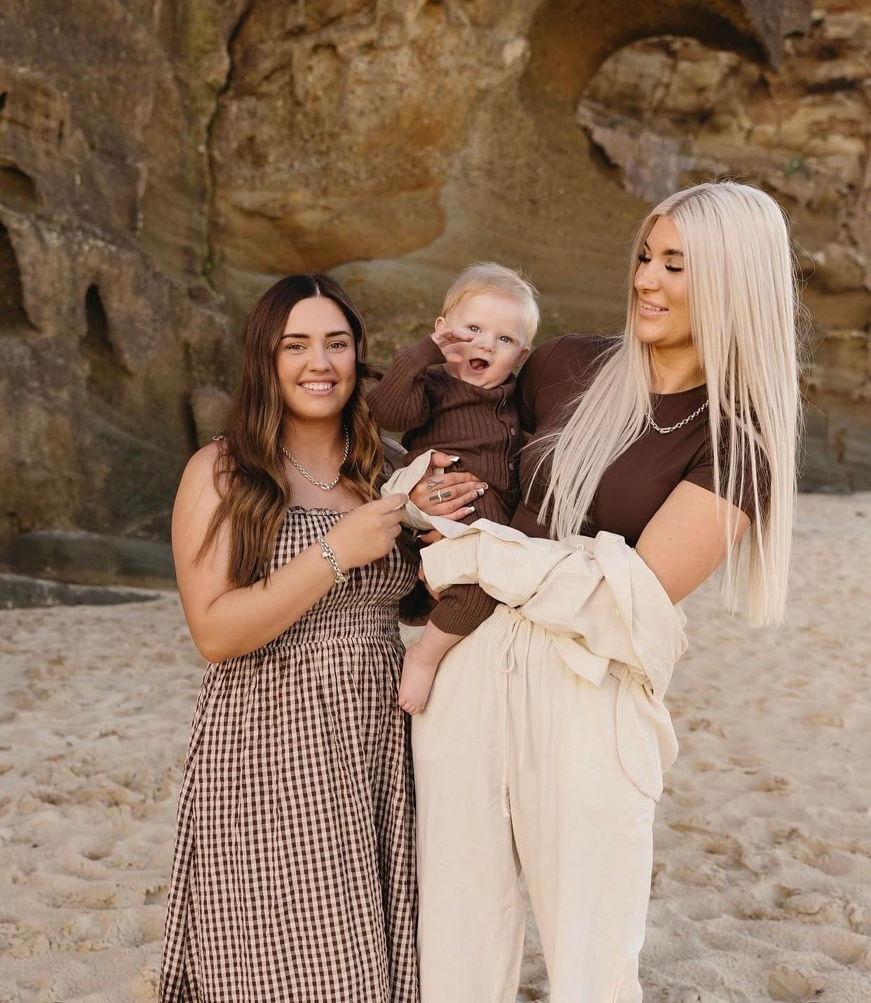 Two women and a baby in brown outfit posing together on a sandy beach with rock formations.