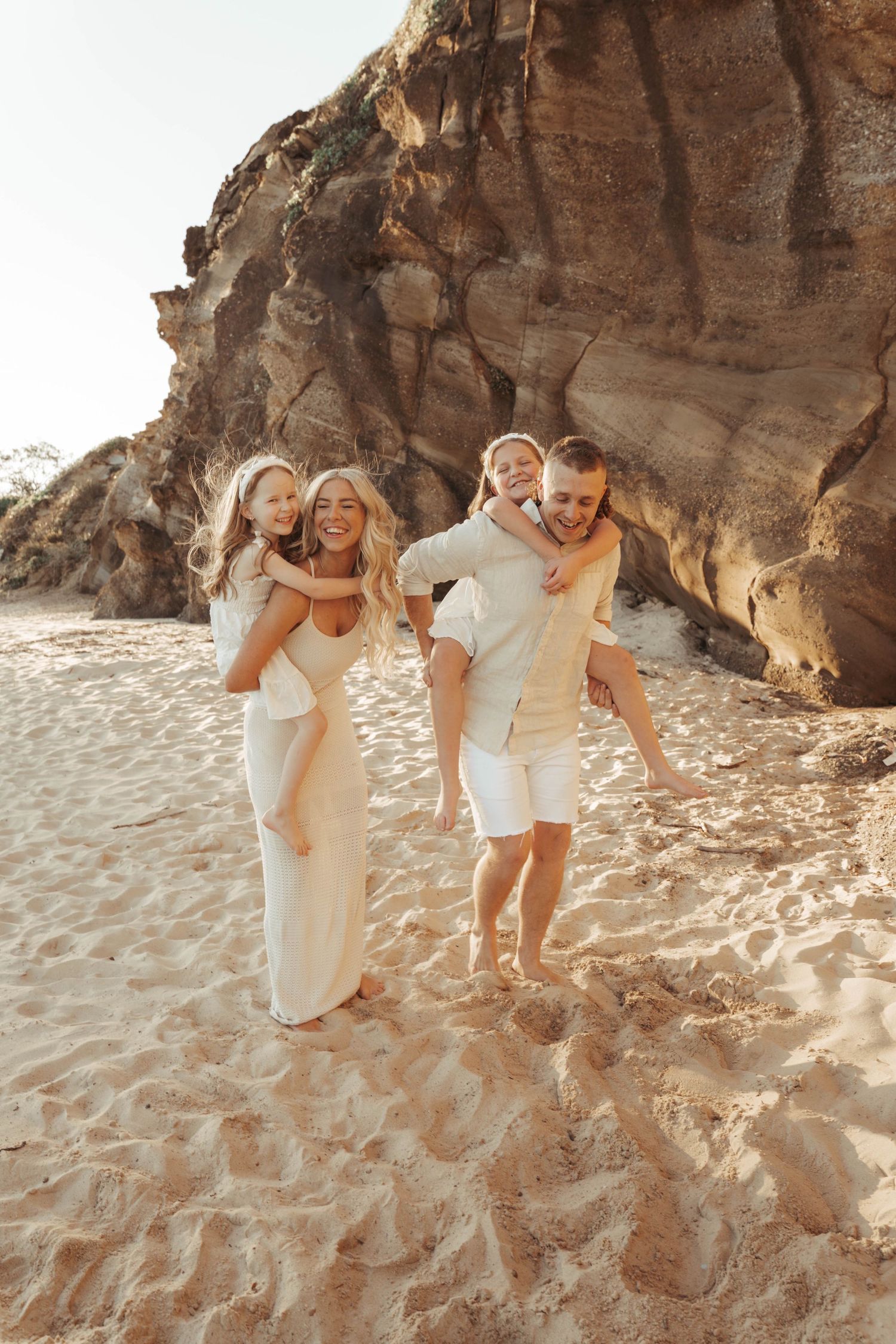 Happy family of four enjoying a sunny beach day, parents giving piggyback rides to children near sandy cliffs.