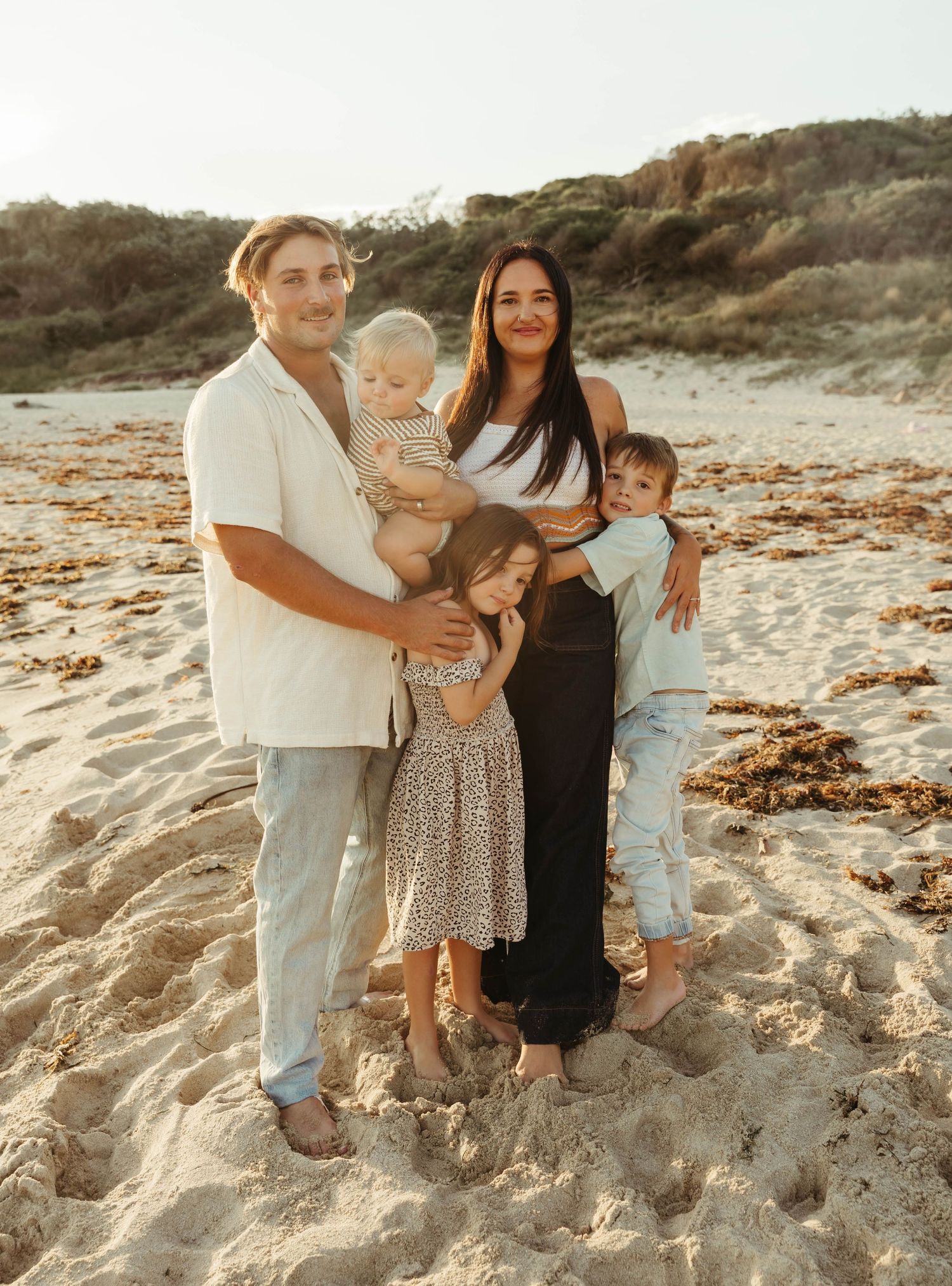 Family of five posing together on a sandy beach at golden hour, parents holding young children with dunes behind.