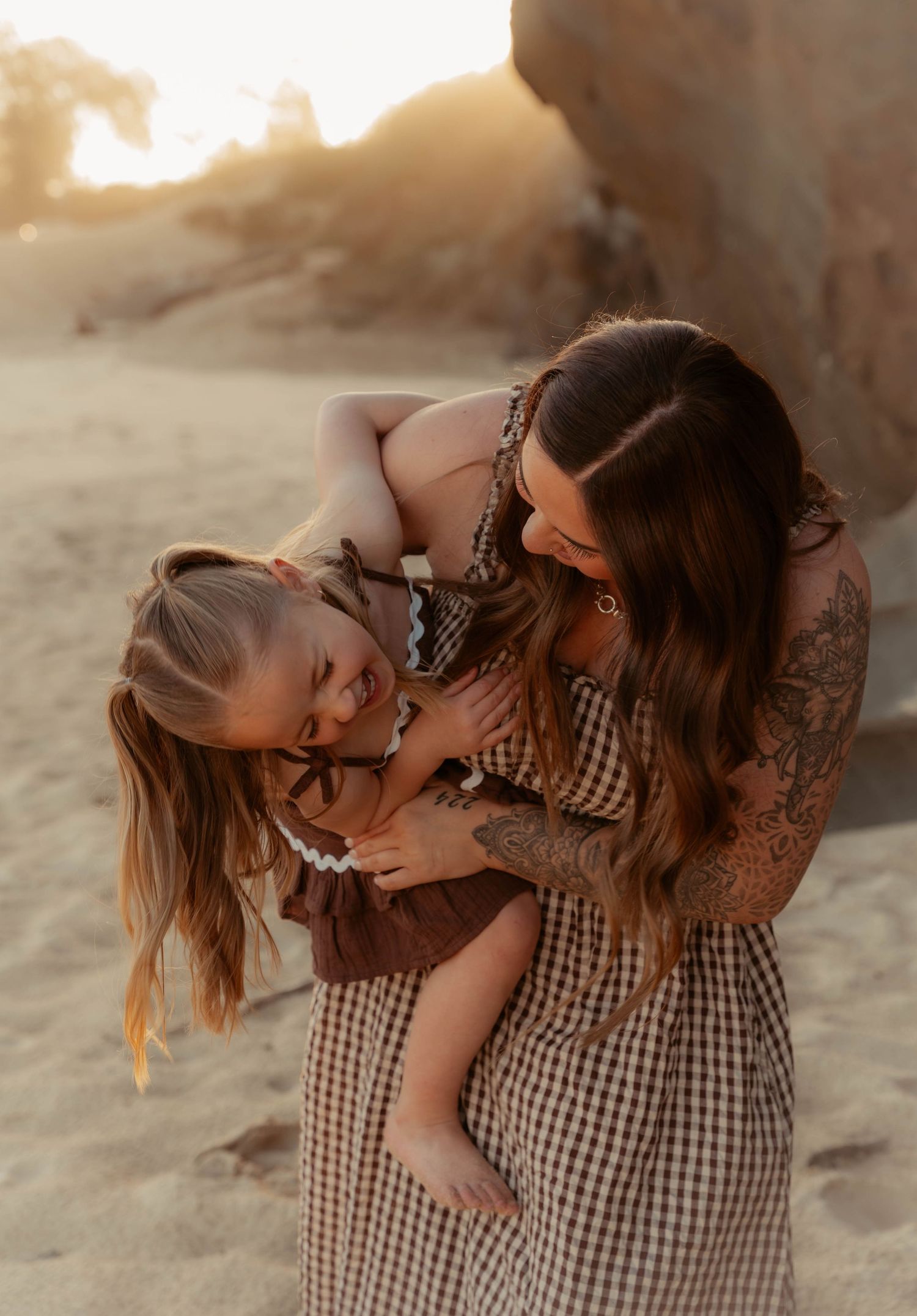 Mother hugging laughing daughter in matching brown gingham dresses on sandy beach.