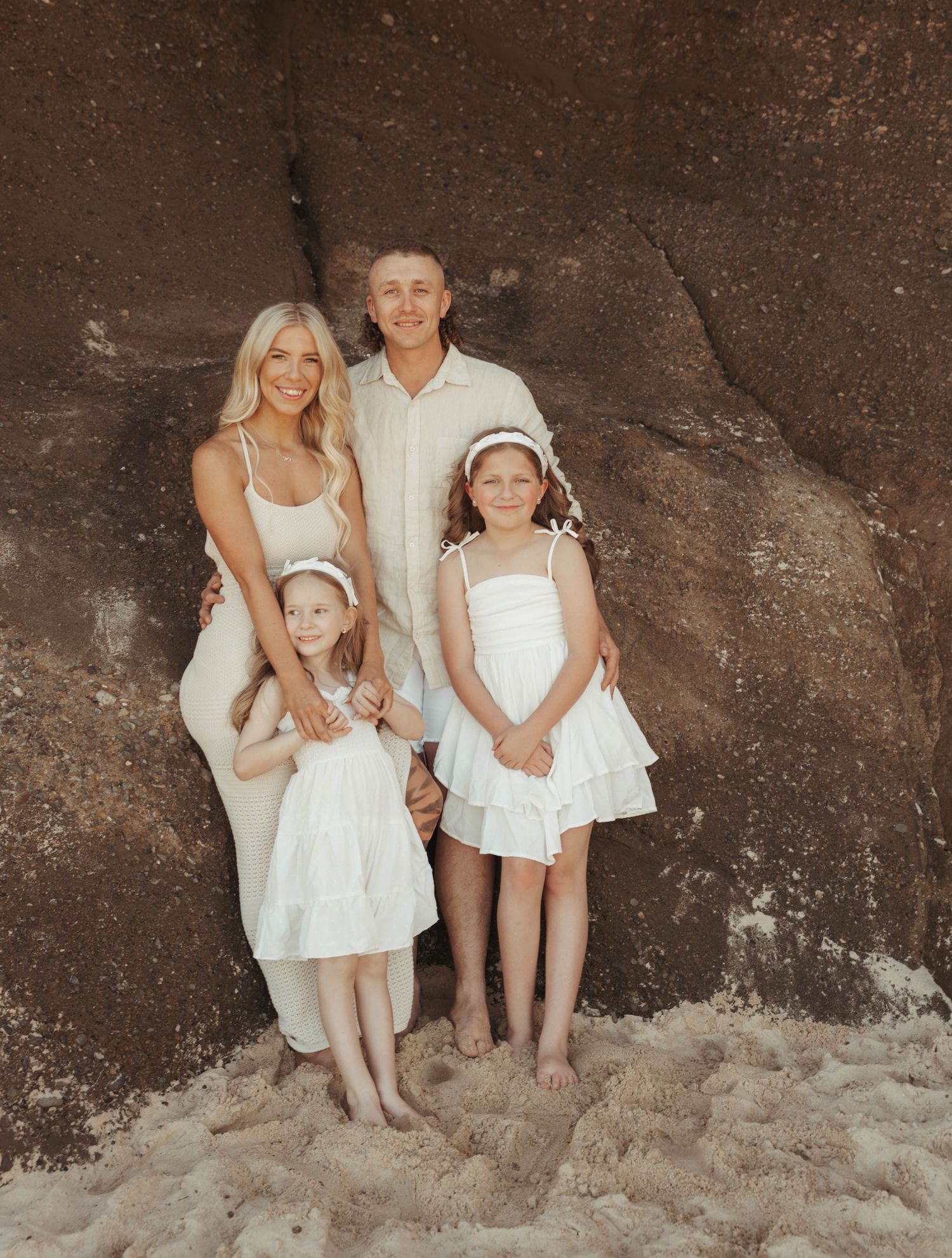 Family of four in white outfits posing together against large brown rock formation on a beach.