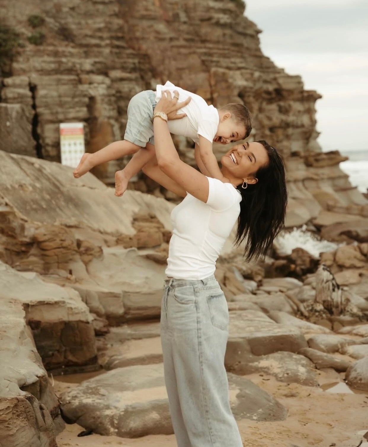 Joyful woman lifting smiling baby overhead near rocky coastal cliffs on a sunny day.