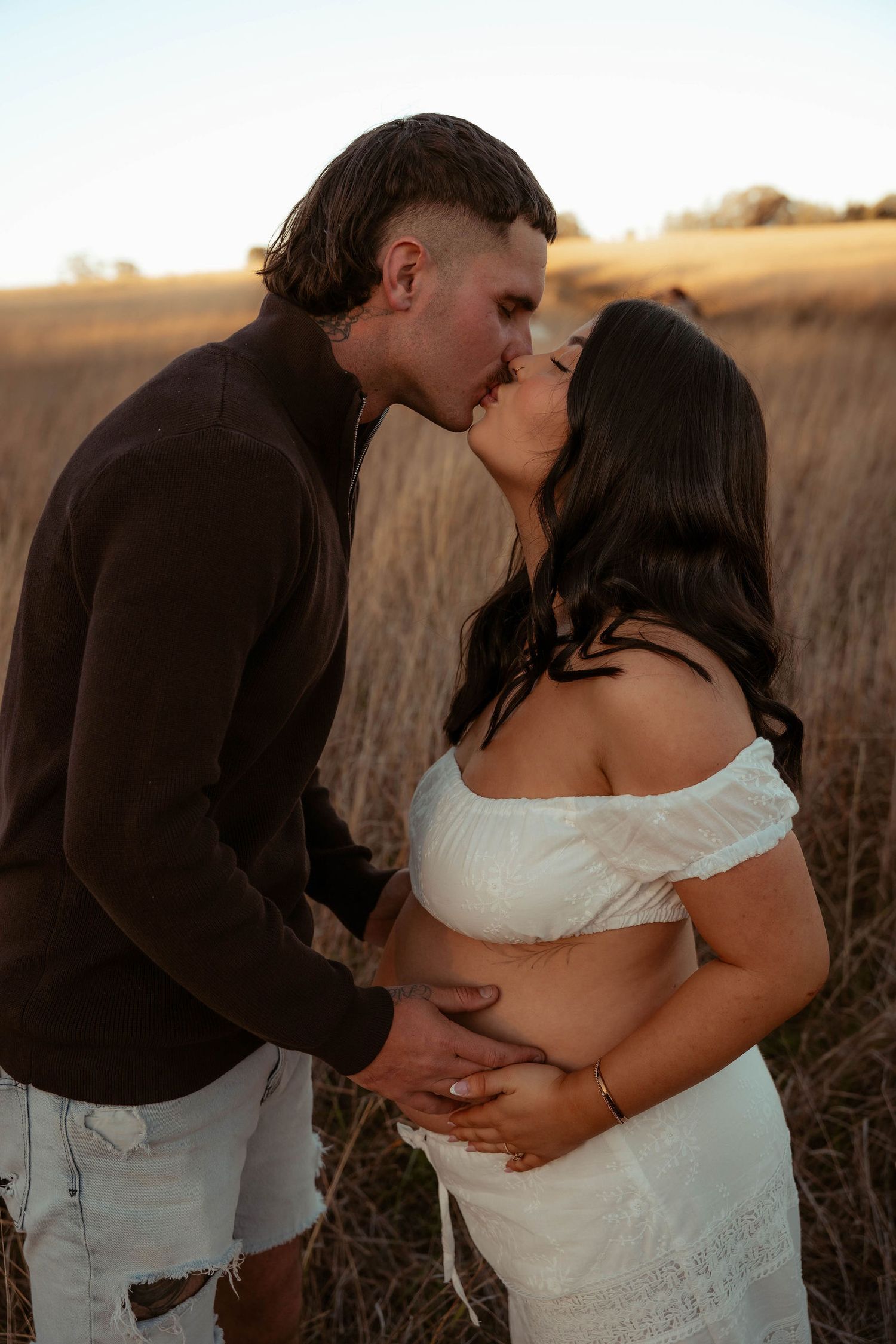 Couple sharing a tender kiss in a golden field at sunset, woman in white off-shoulder dress cradling baby bump.